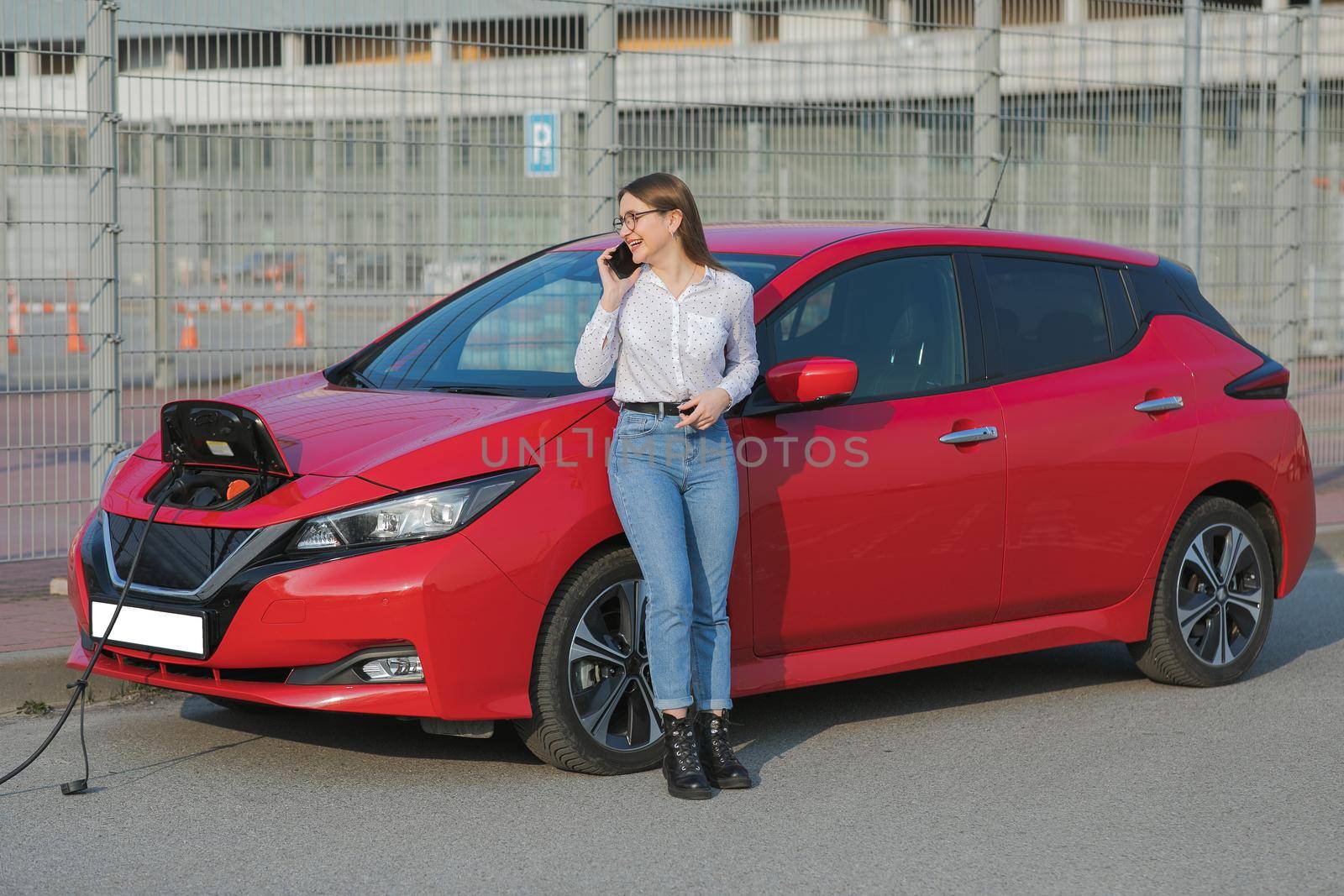 Girl stands with phone near her red electric car and waits when vehicle will charged. Plugging in power cord to an electric car. Ecological car connected and charging batteries by uflypro