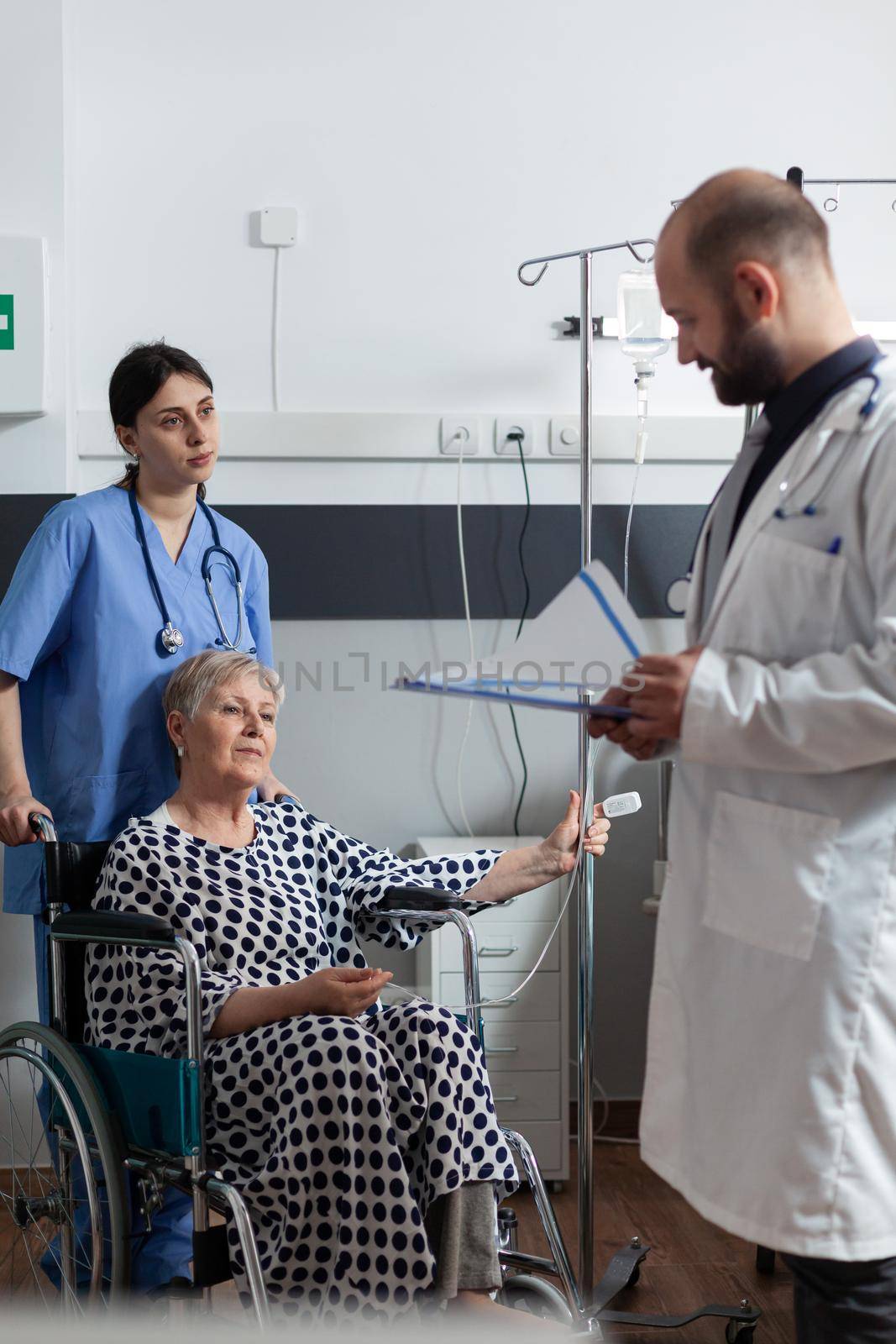 Elderly sick patient sitting in hospital wheelchair getting treatment through iv drip bag and oxymeter attached on finger monitoring oxygen saturation from blood. Doctor discussing about recovery therapy.