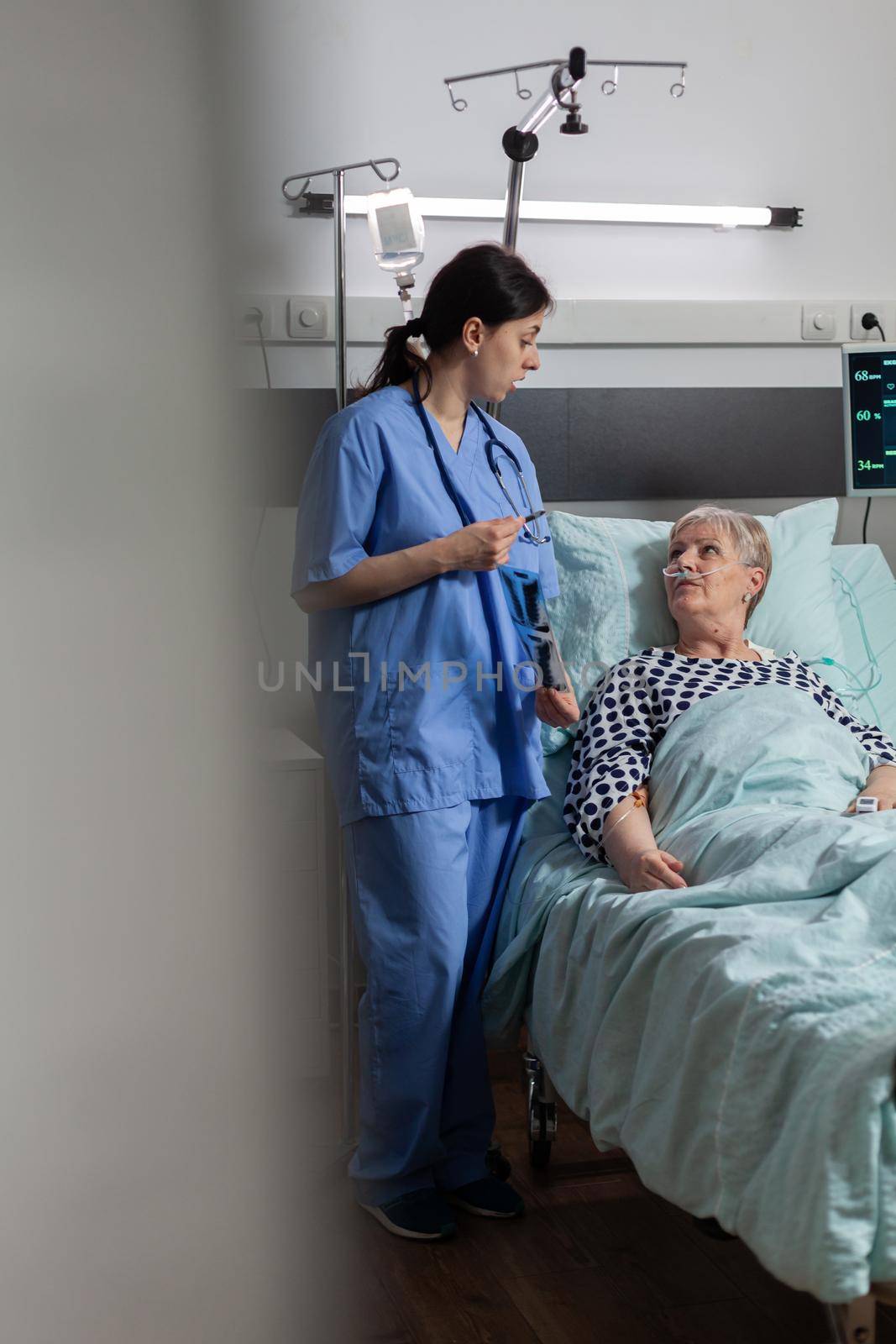 Nurse discussing with elderly patient laying in hospital bed during visit about chest x-ray. Recieving treatment through intravenous line and inhale and exhale with help from oxygen mask.