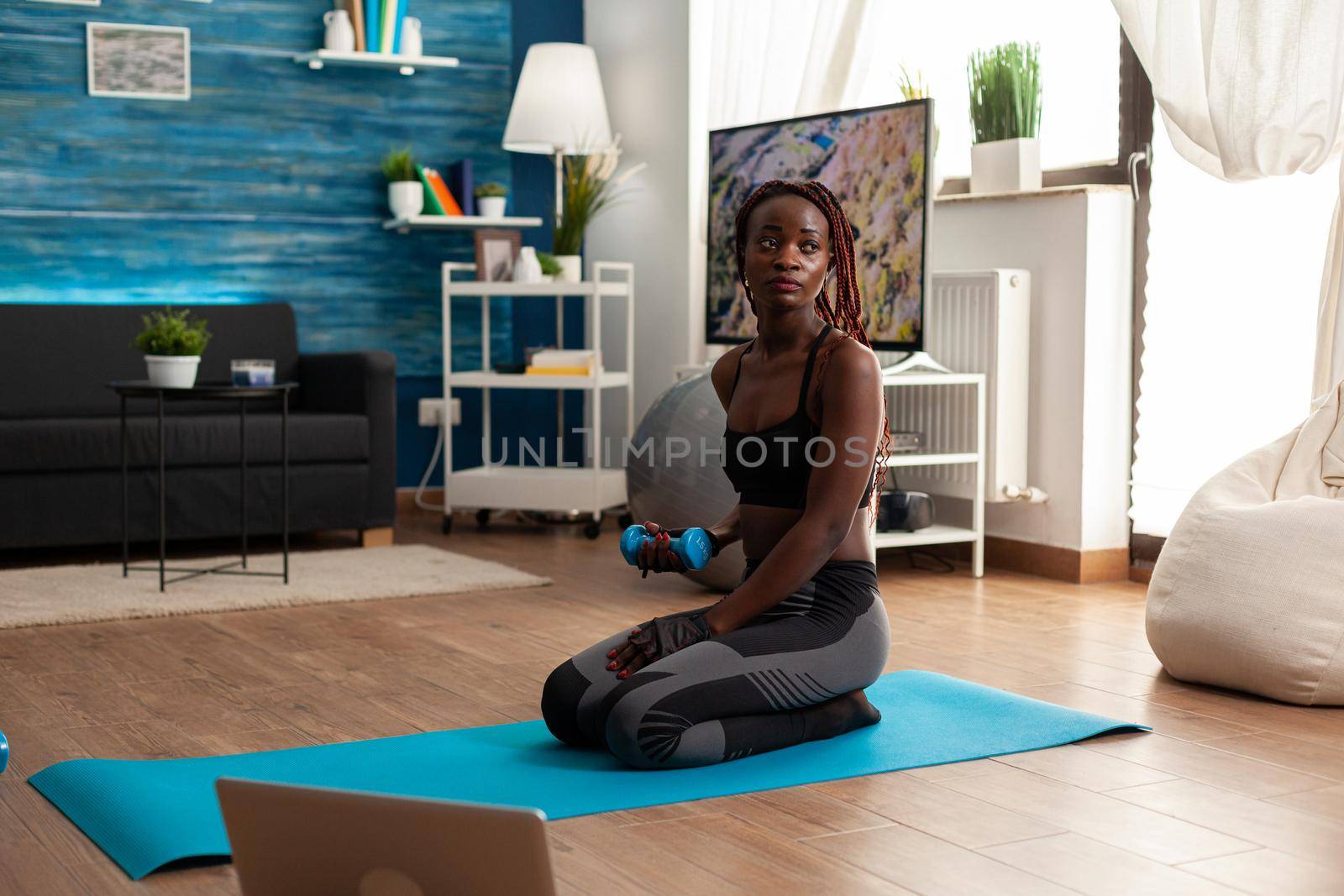 Fit athletic active black woman flexing arms working biceps, sitting on yoga mat in home living room exercising following video instructions from laptop, having a healthy lifestyle.