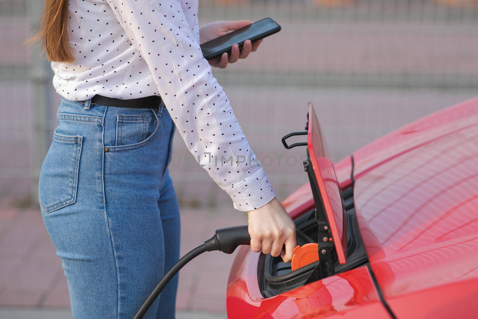 Caucasian girl using smart phone and waiting power supply connect to electric vehicles. Process of car's electrical recharge comes to end. by uflypro