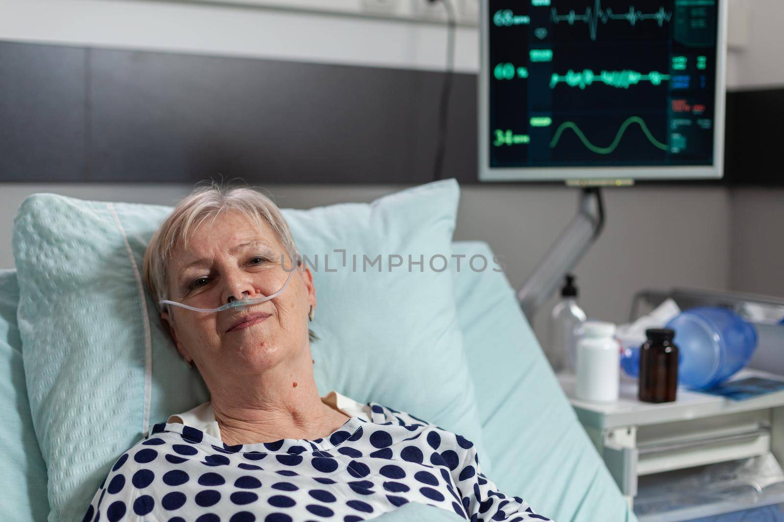Senior woman patient laying in hospital bed following recovery treatment, getting receiving a medication through an intravenous line. Oxymeter attached on finger monitoring oxygen blood saturation.