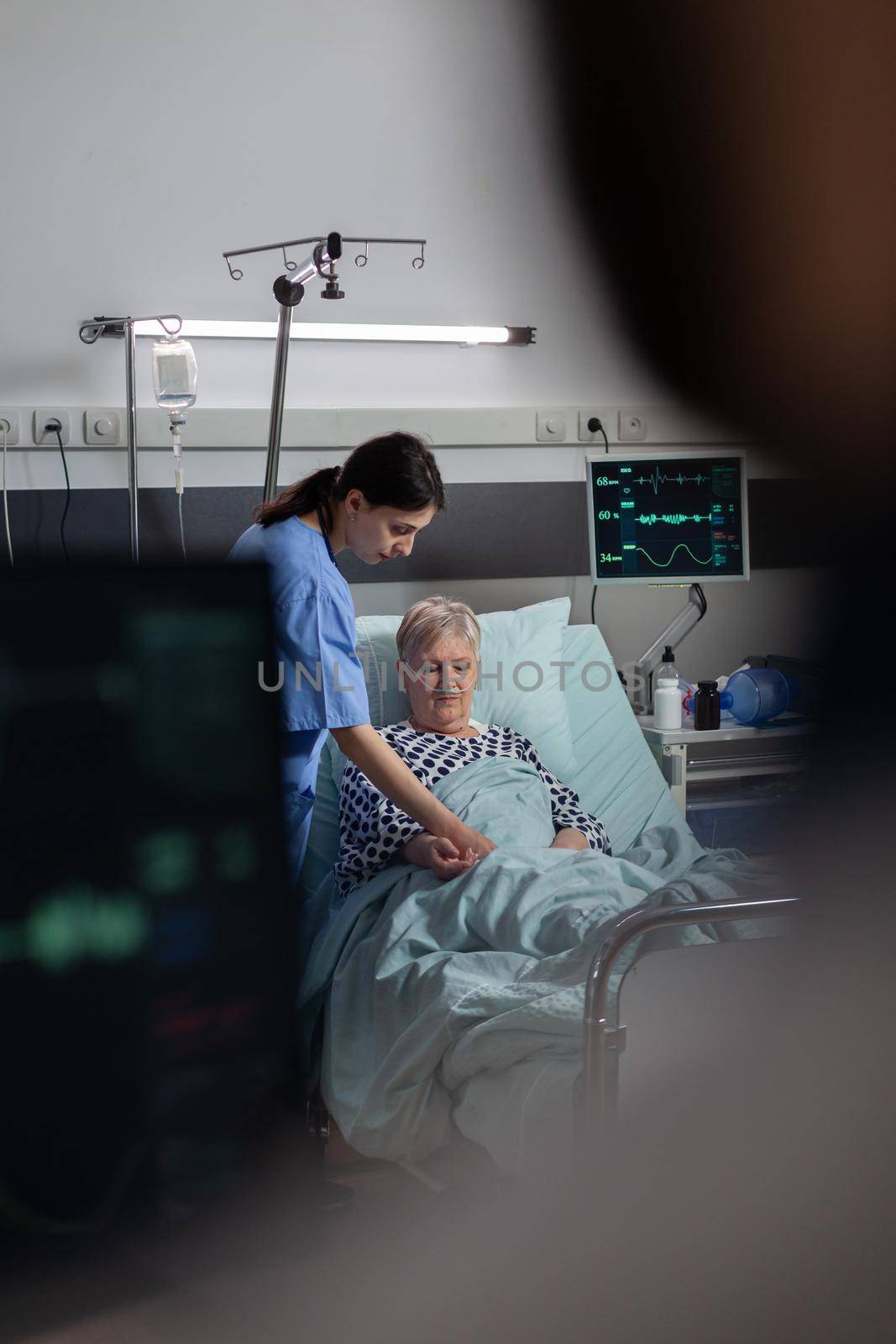 Friendly doctor reassuring sick old patient holding hands, showing emphaty, encouragement, during medical examination in hospital room. Patient breathing through oxygen mask.