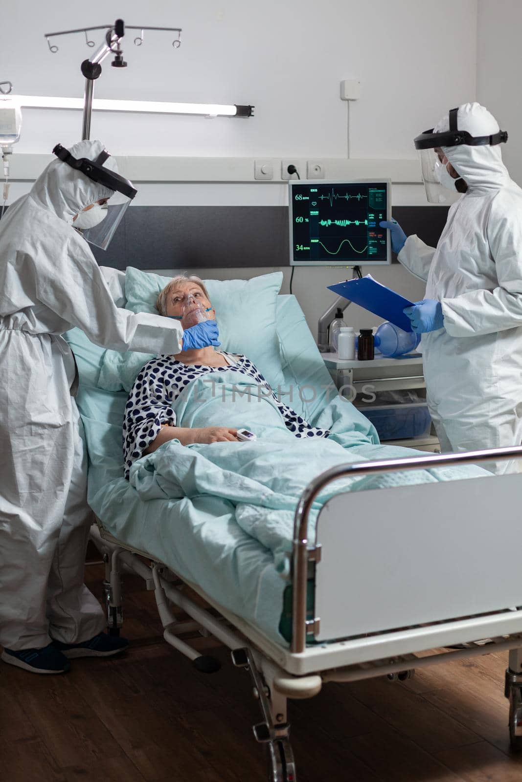 Doctor and nurse wearing cover all ppe suit as prevention for infection with coronavirus during medical vizit at senior patient, in hospital room who breaths with oxygen mask.