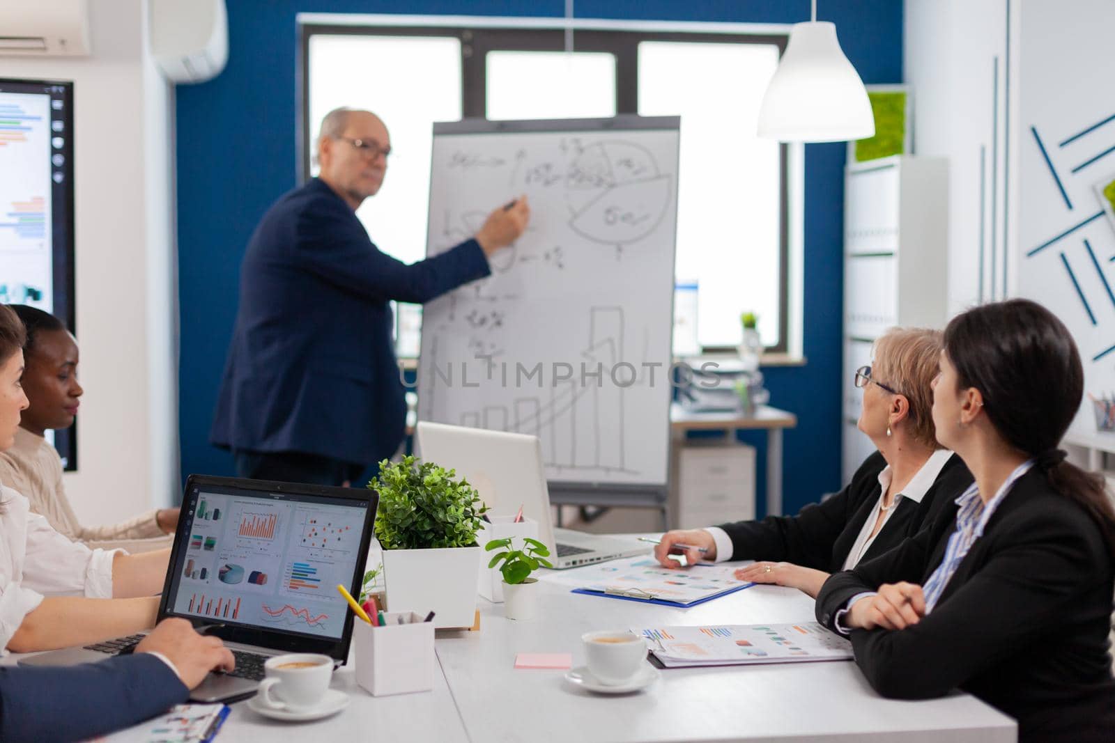 Elderly businessman giving presentation to multiethnic team of colleagues using flip chart. Serious speaker boss executive, business trainer explaining development strategy to motivated mixed race employees.