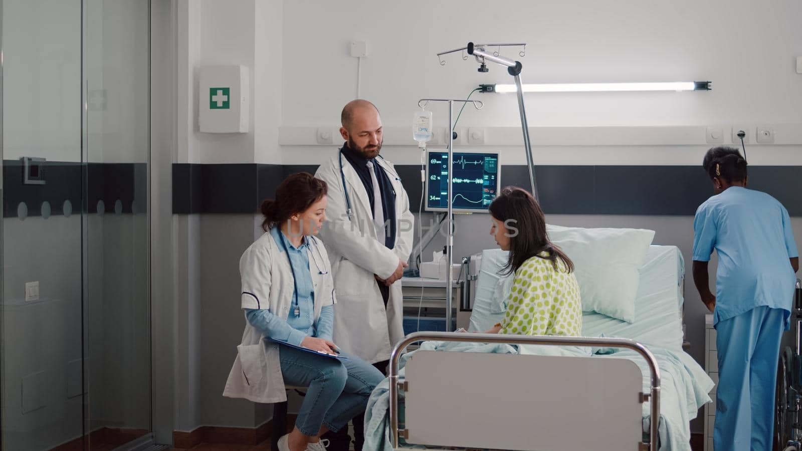 Specialist practitioner doctors monitoring sick man explaining illness treatment while working in hospital ward. Patient sitting in bed discussing symptom disease during recovery appointment