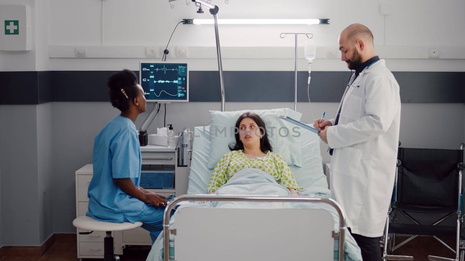 Sick woman lying on bed while afro-american nurse analyzing x-ray bones monitoring disease symptoms. Surgeon man consulting recovery patient writing ill treatment on clipboard in hospital ward