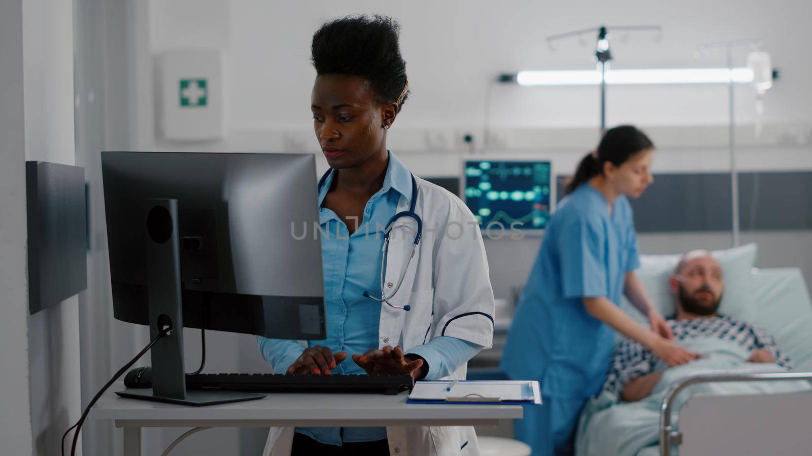 Front view of afro american doctor typing medical expertise on computer while in background nurse checking heartbeat pulse using oximeter. Hospitalized patient having respiratory disorder