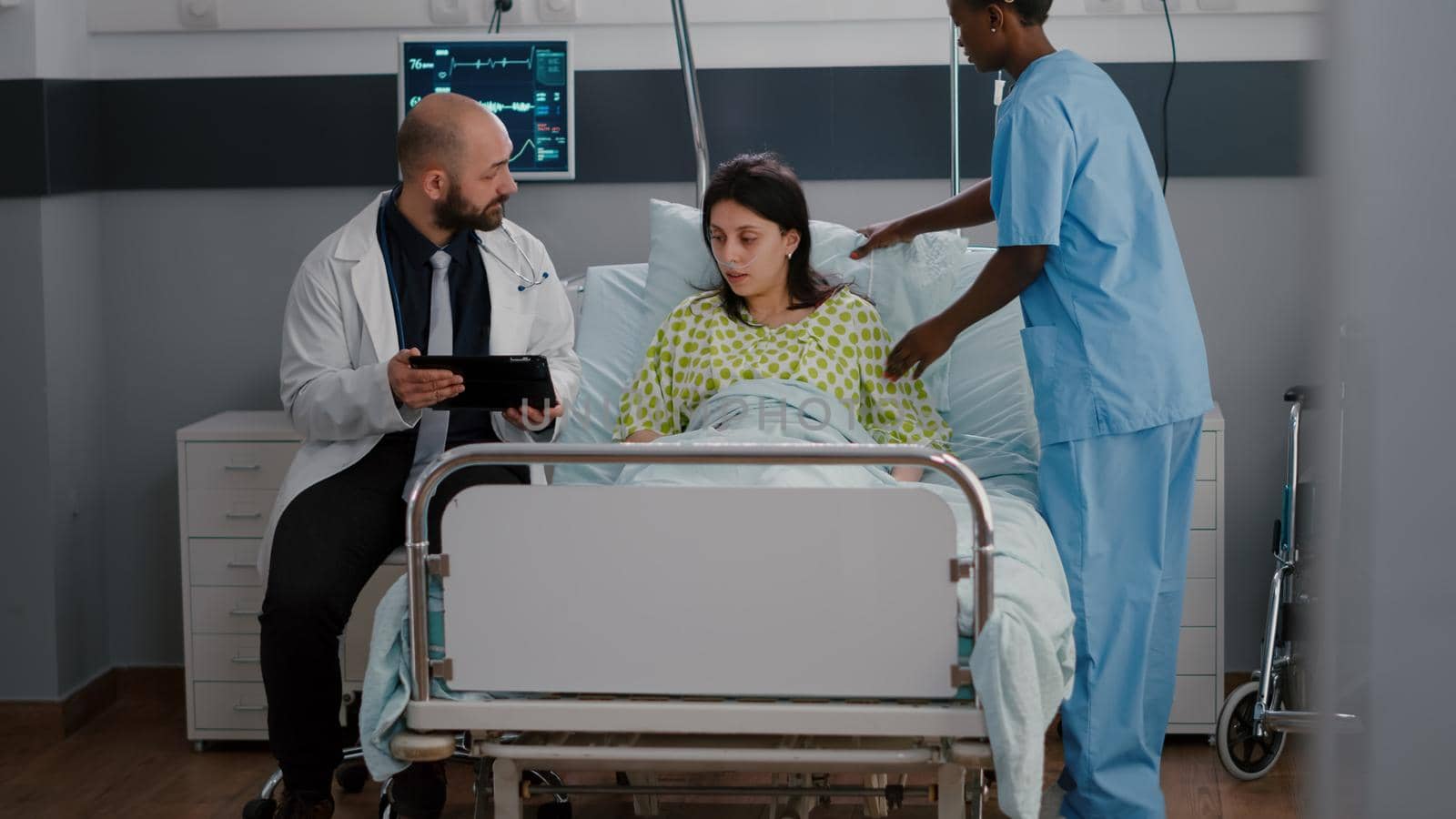 Practitioner physician doctor explaining disease expertise to sick woman using tablet computer. Patient sitting in bed recovering after medical surgery in hospital ward while black nurse arrange bed