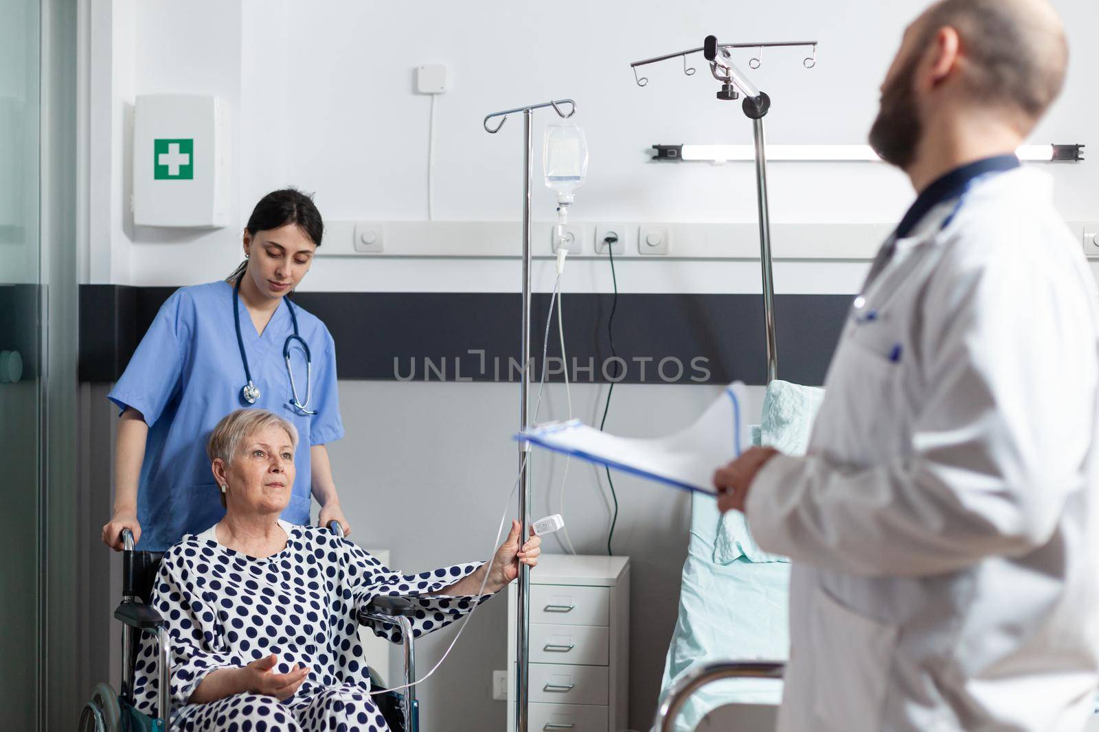 Sick senior woman hospitalized sitting in wheel chair with iv drip bag attached on arm, helped by nurse to move. Doctor with stethoscope checking patient before surgery and talking about treatment recovery.