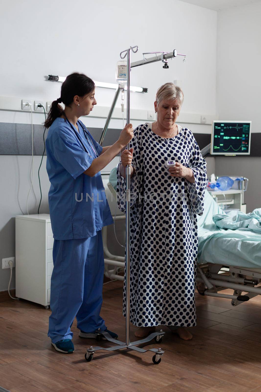 Sick senior woman carrying iv drip bag with help from medical nurse by DCStudio