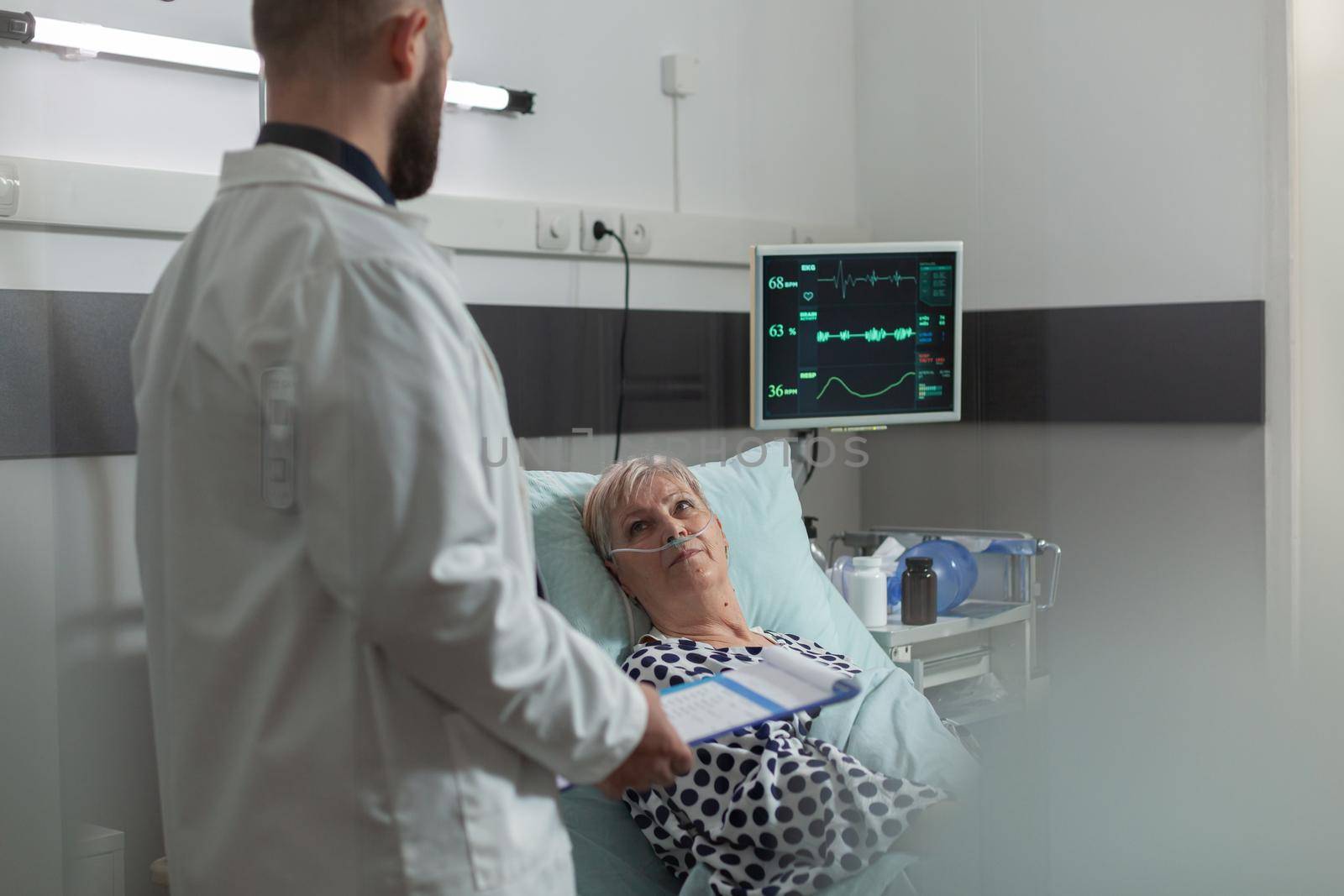 Doctor reassuring senior woman full recovery during discussion consultation in hospital room. Writing note on clipboard. Breathing with help from oxygen mask.