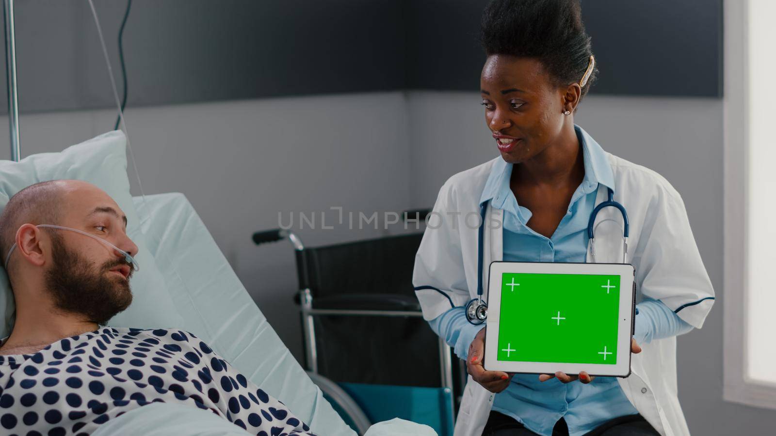Doctor with dark skin explaining to woman patient resting in bed about sickness treatment. Practitioner holding mock up green screen chroma key tablet computer with isolated display in hospital ward