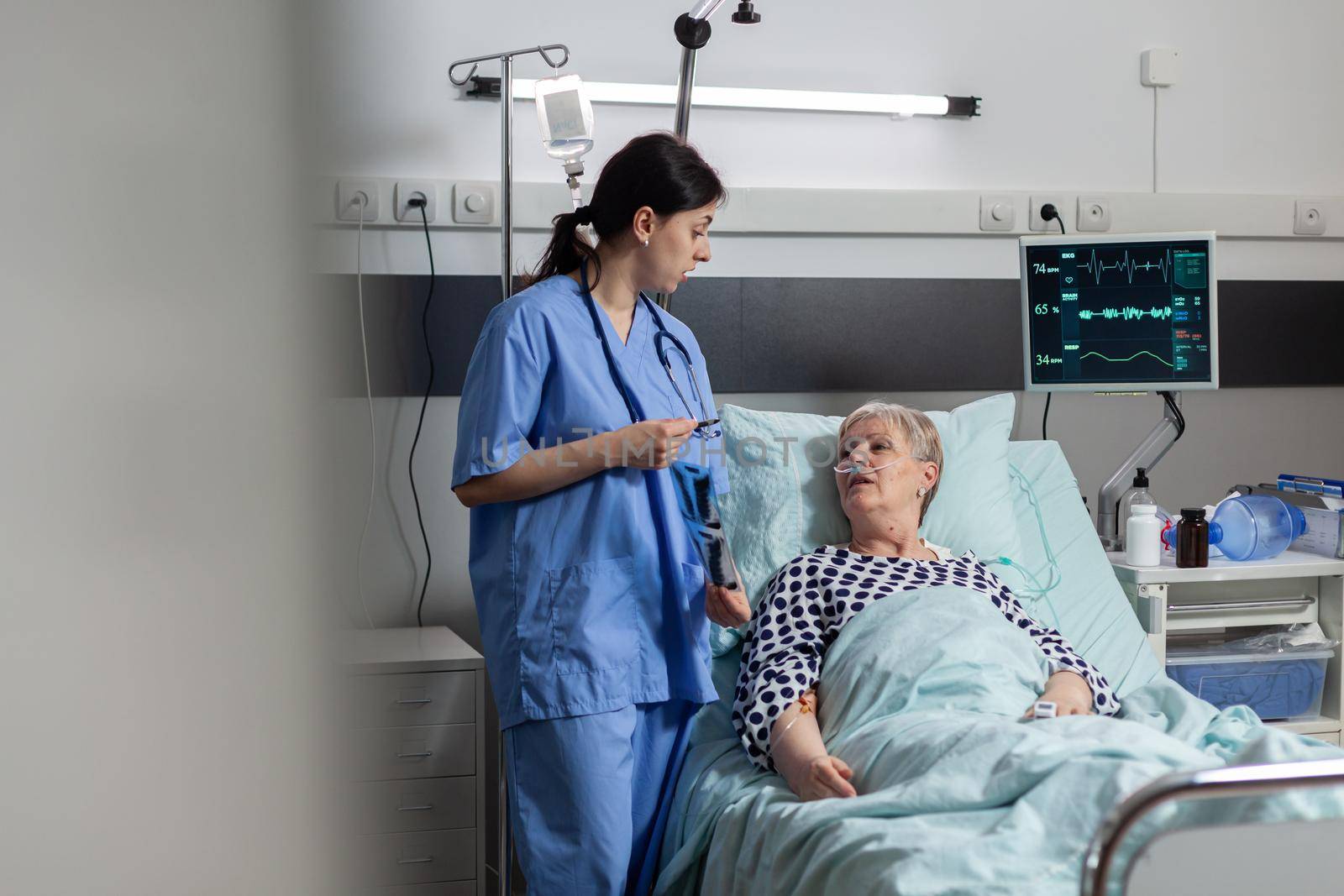 Nurse discussing with elderly patient laying in hospital bed during visit about chest x-ray. Recieving treatment through intravenous line and inhale and exhale with help from oxygen mask.