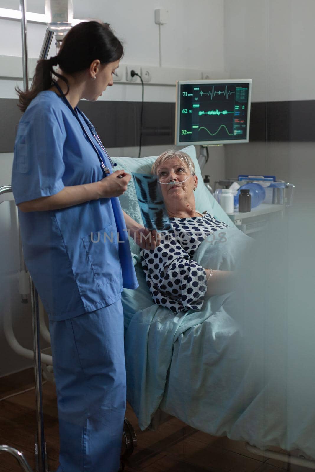 Elderly patient in radiology hospital room laying in bed listening nurse dicussing about chest x-ray radiography for respiratory diagnosis. Breathing with help from oxygen mask.