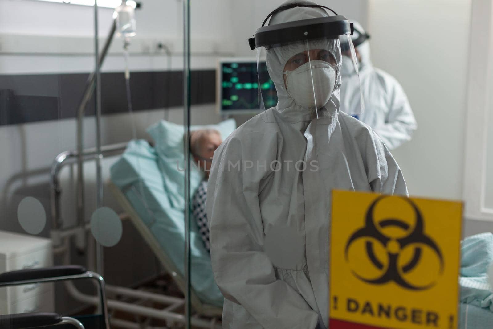 Portrait of medical nurse dressed in pee suit in hospital room looking at camera, with senior woman patient laying in bed breathing heavely with help from oxygen mask.