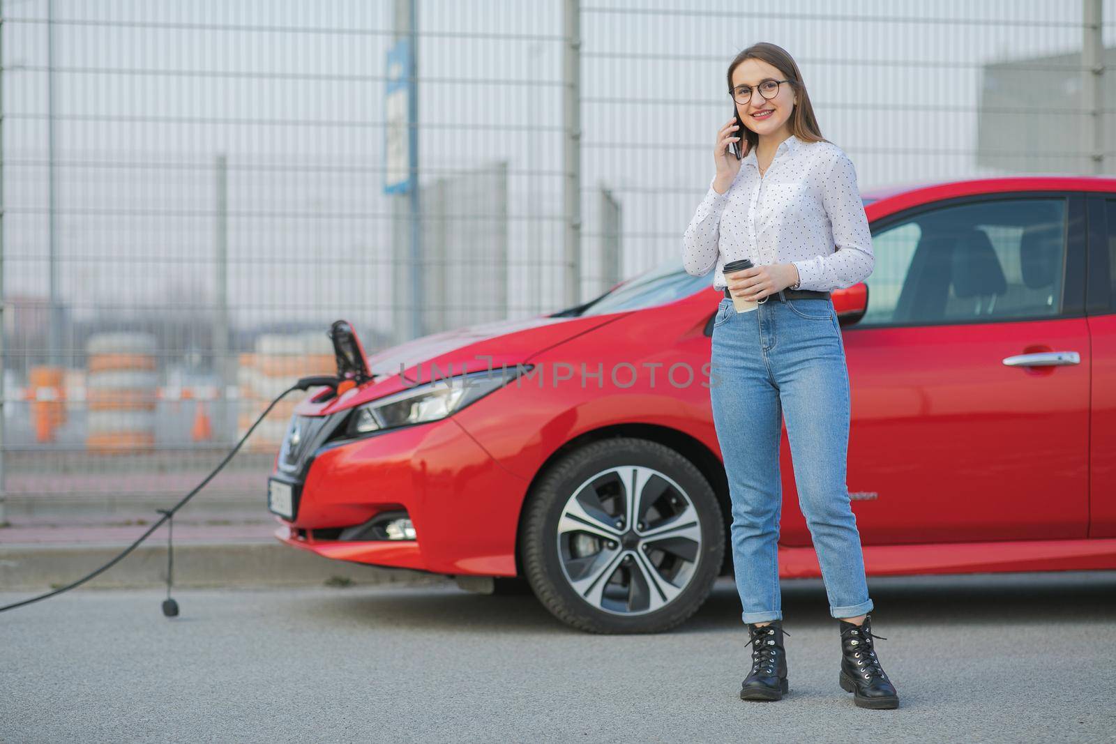 Electric car charging in street. Ecological Car Connected and Charging Batteries. Girl Use Coffee Drink While Using SmartPhone and Waiting Power Supply Connect to Electric Vehicles for Charging.