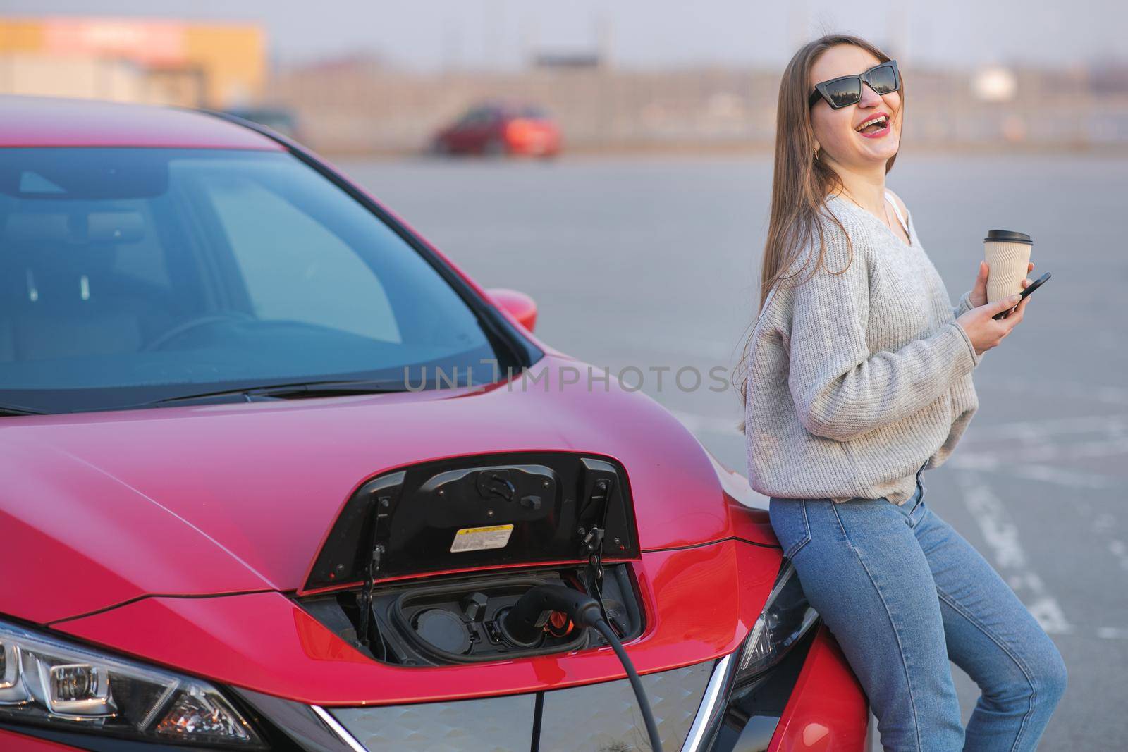 Girl Use Coffee Drink While Using Smart Phone and Waiting Power Supply Connect to Electric Vehicles for Charging the Battery in Car. by uflypro