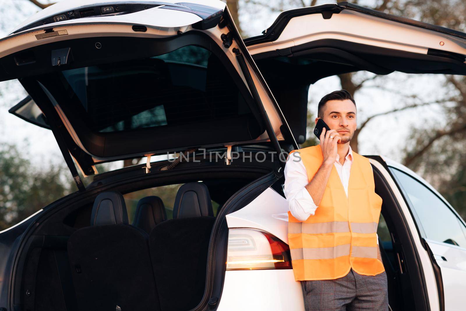 Man with reflective vest calling by phone about breakdown in her electric car. Emergency stop sign. Electric car accident on the road by uflypro