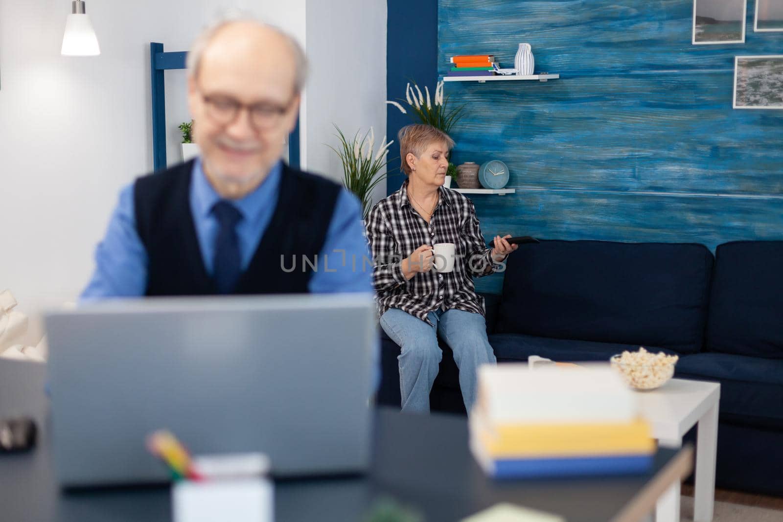 Old woman relaxing on couch holding a cup of coffee and husband is working. Elderly man entrepreneur in home workplace using portable computer sitting at desk while wife is holding tv remote.