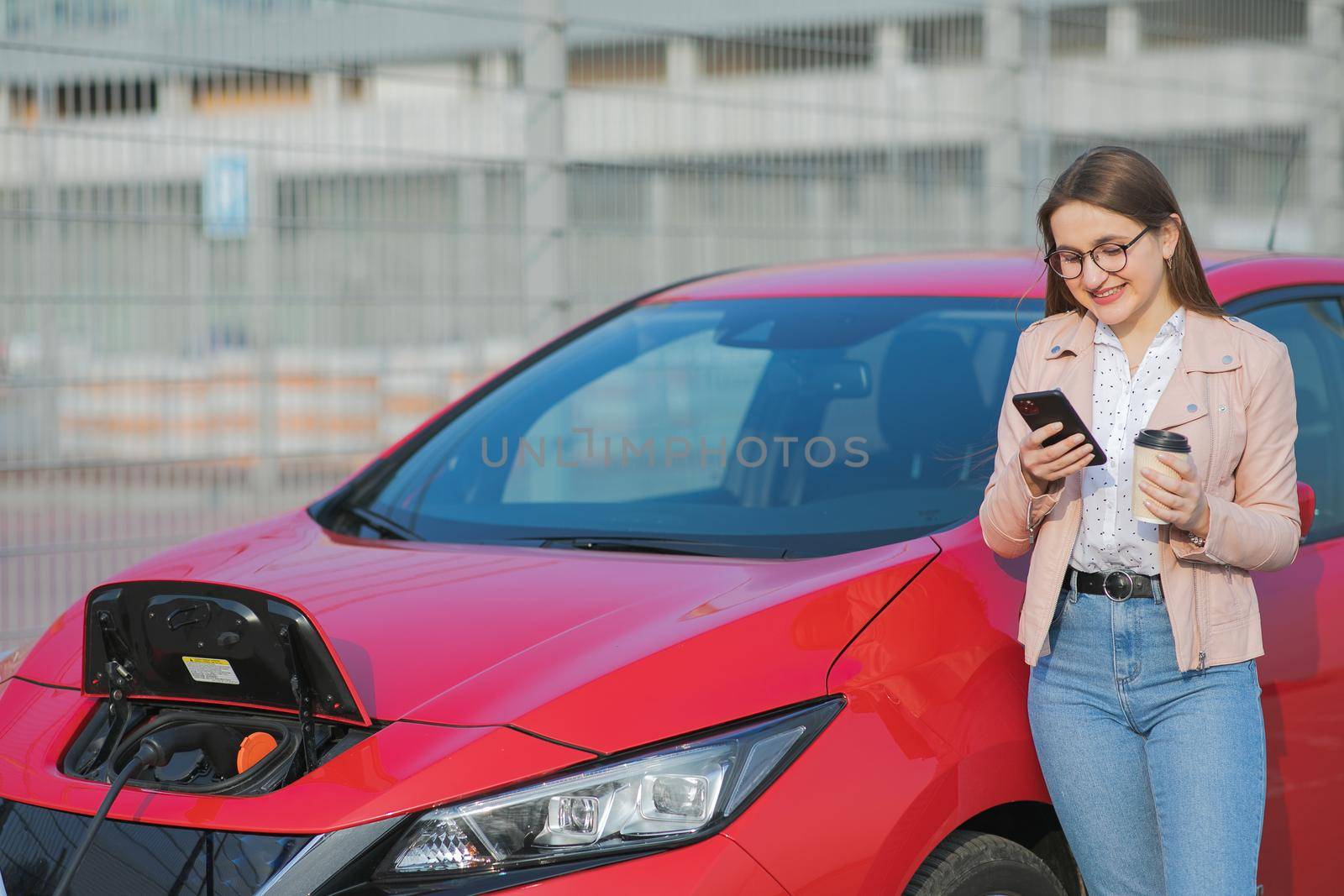 Girl Use Coffee Drink While Using Smart Phone and Waiting Power Supply Connect to Electric Vehicles for Charging the Battery in Car.