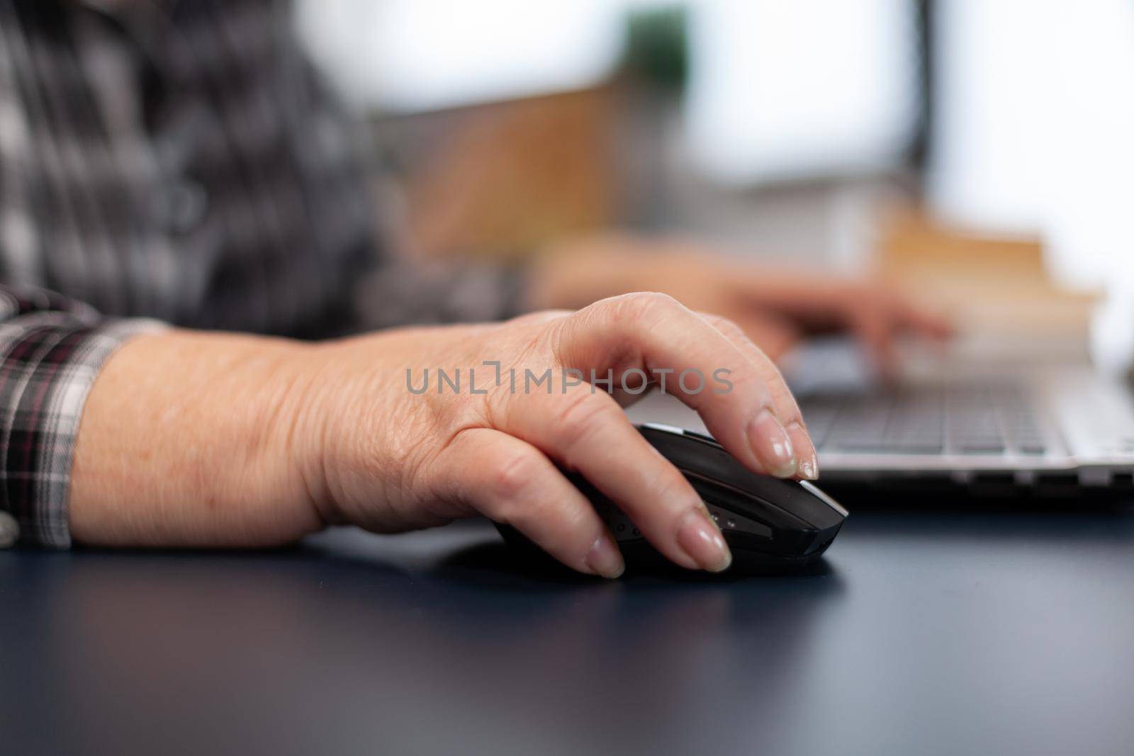 Close up of old entrepreneur lady using mouse in home office. Elderly woman in home living room using moder technoloy laptop for communication sitting at desk indoors.