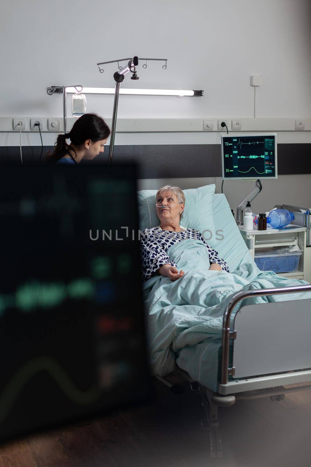 Medical nurse in scrubs showing compassion to senior woman patient, laying in hospital bed breathing with help from oxygen mask, getting intravenous medicine from iv drip bag.