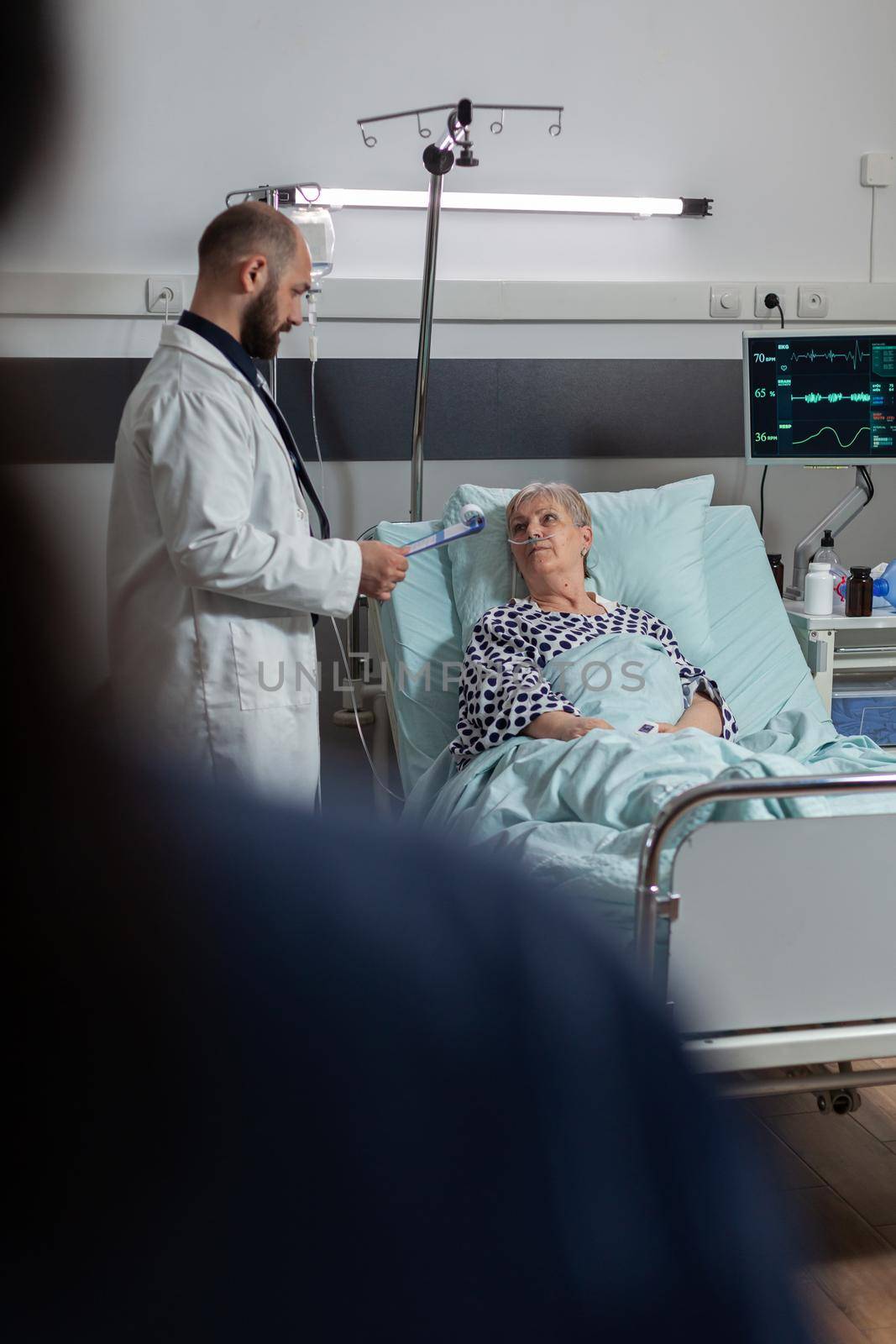 Doctor questioning senior woman laying in hospital bed, breathing with help from oxygen tube because of lungs surgery. Patient recieving medicine through intrevenous line.