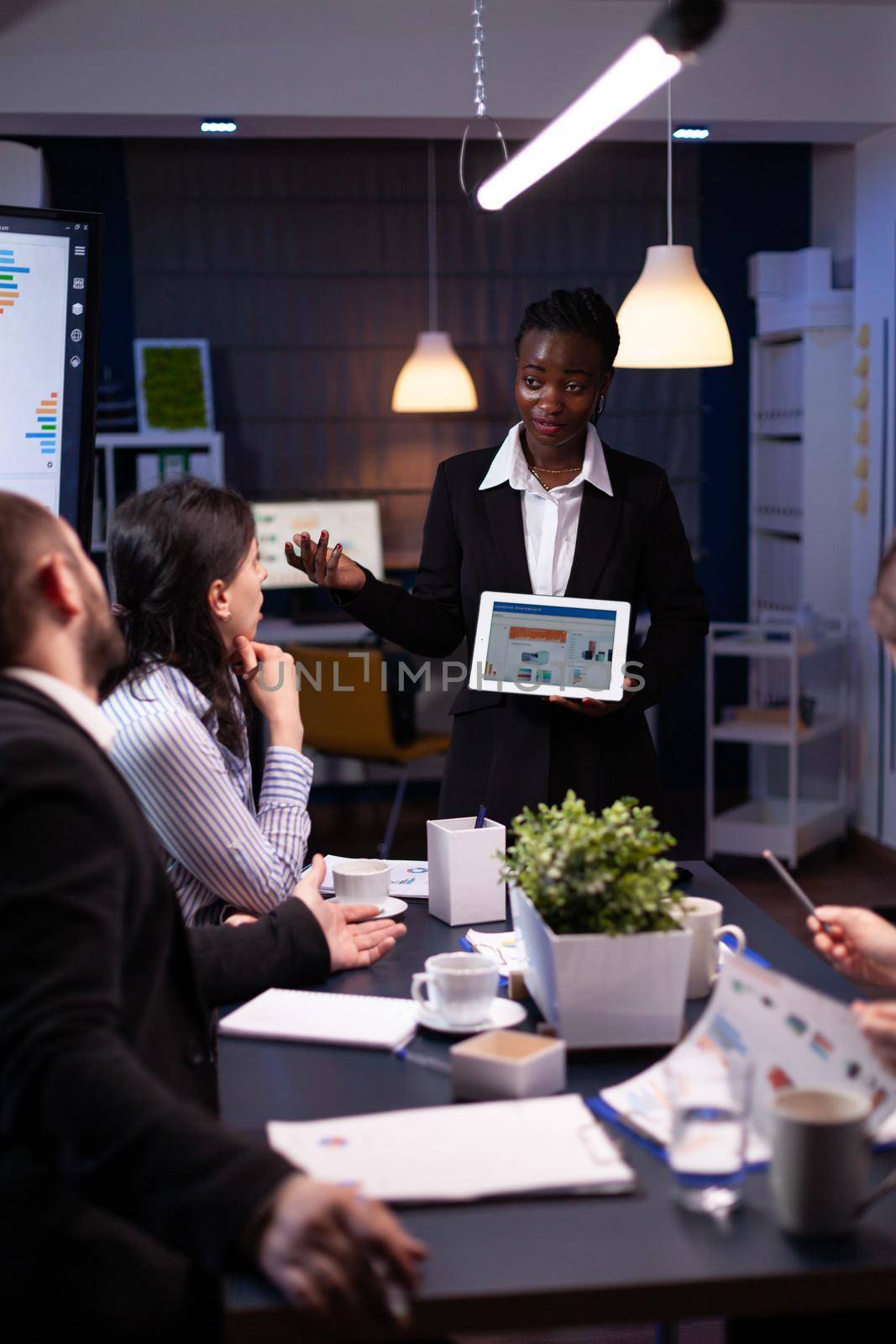 Entrepreneur woman with dark skin overworking in company office meeting room explaining management strategy using tablet. Workaholics multi-ethnic businesspeople brainstorming late at night.
