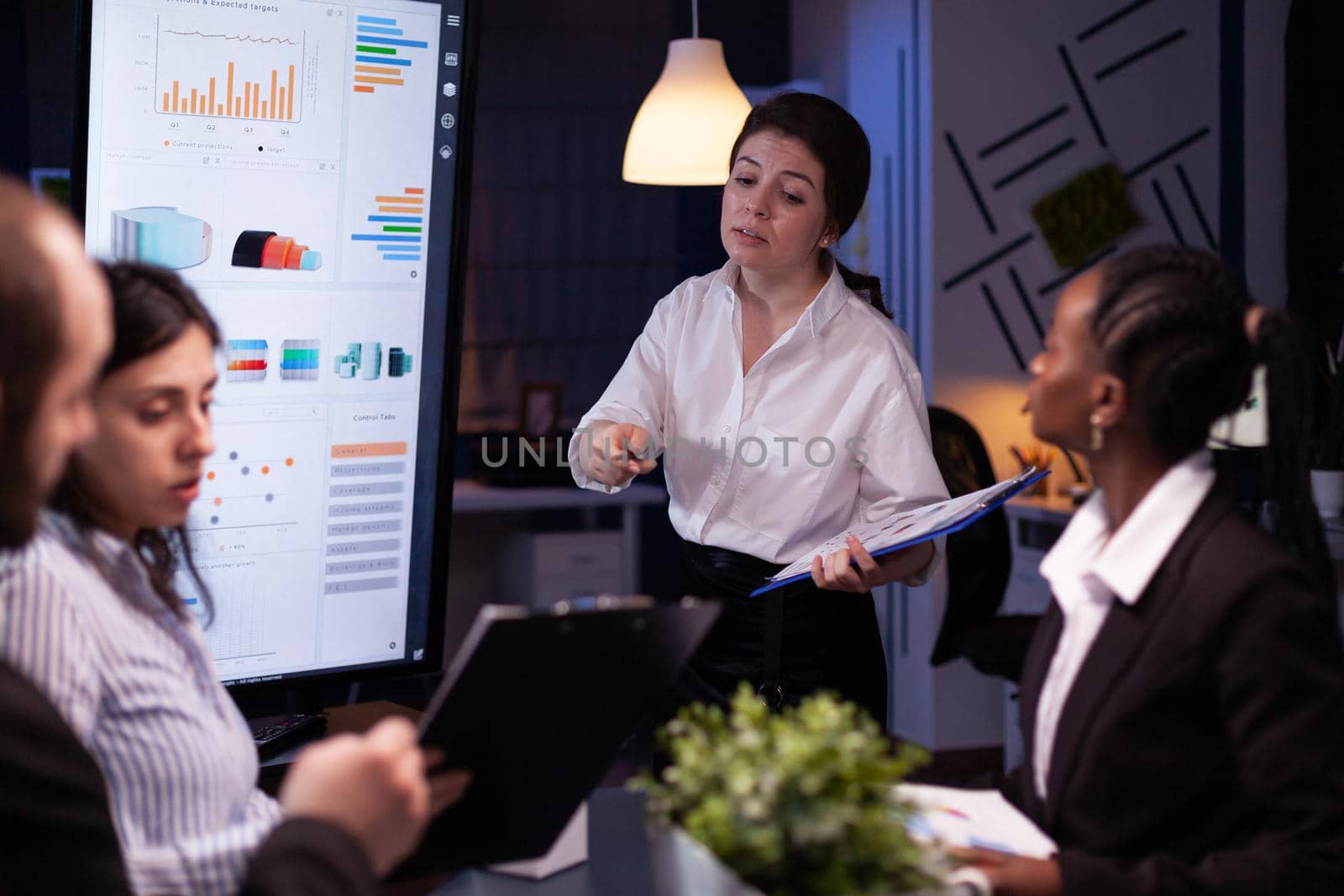 Diverse multiethnic business teamwork overworking in office meeting room analyzing financial graphs late at night. Coworkers brainstorming project ideas solving company problem.