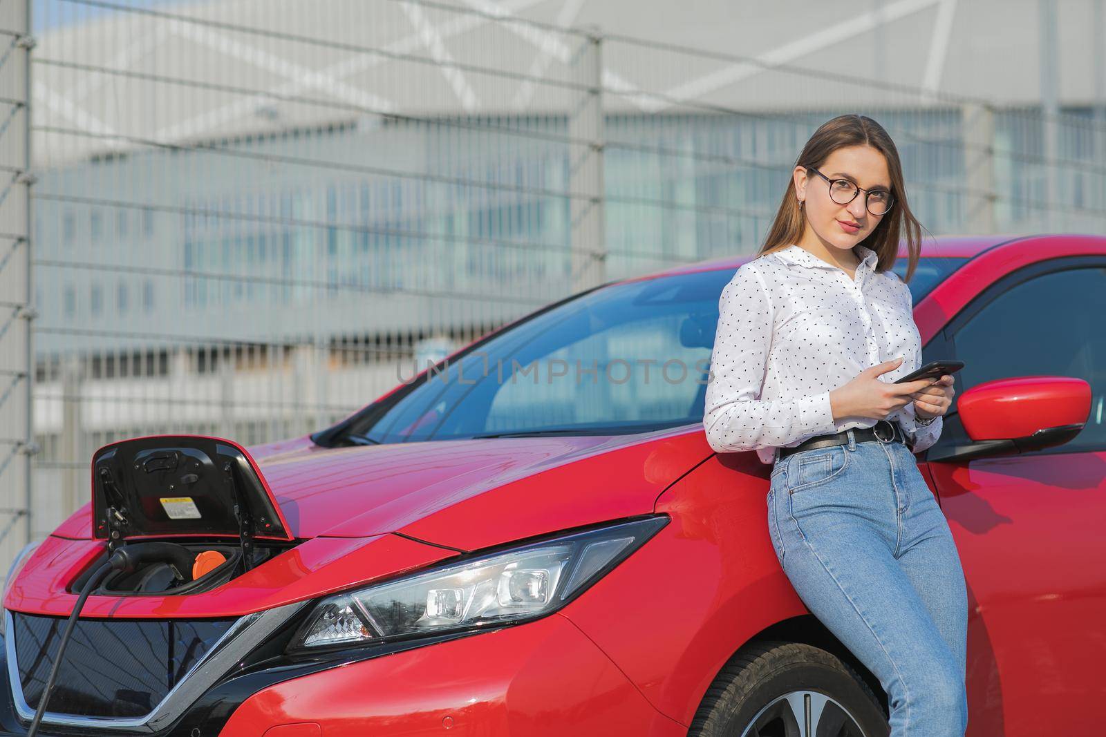 Girl using smart phone and waiting power supply connect to electric vehicles for charging the battery in car. Ecological car connected and charging batteries by uflypro