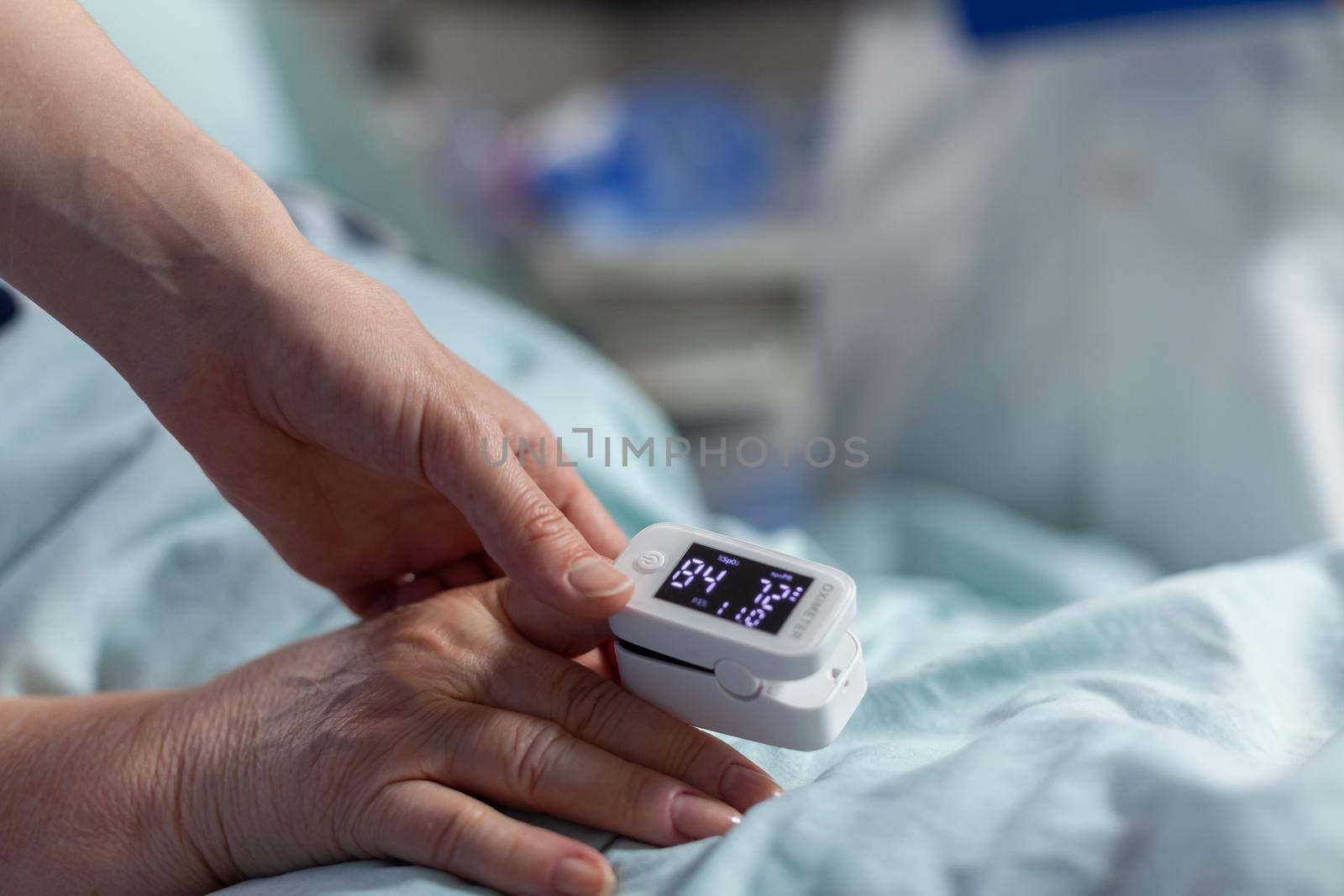 Close up of oxymeter attached on sick senior patient laying in hospital bed, monitoring hemoglobin. Medical staff using modern technology for reading blood oxygen saturation.