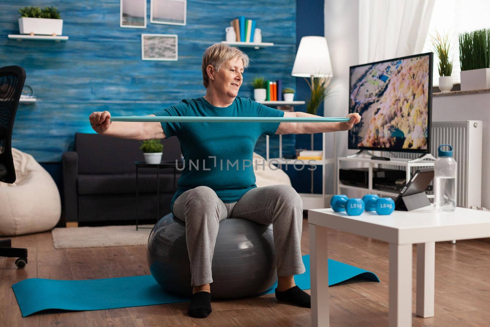 Retired senior man sitting on fitness swiss ball in living room doing wellness fitness workout by DCStudio
