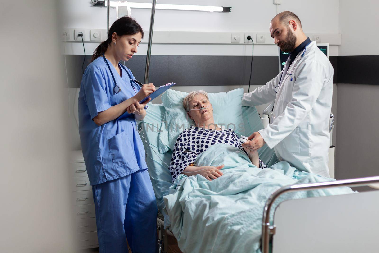 Nurse is taking notes on clipboard during consultation of senior woman, while doctor is reading data from oxymeter about pulse, blood pulse and oxygen saturation attached on sick patient attached on finger.