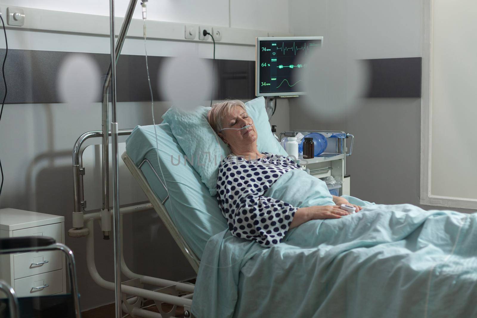 Senior woman patient in hospital room getting medicine through intravenous line from iv drip bag and breathing with help from oxygen mask laying in bed. Oxymeter attached on finger monitoring bpm.