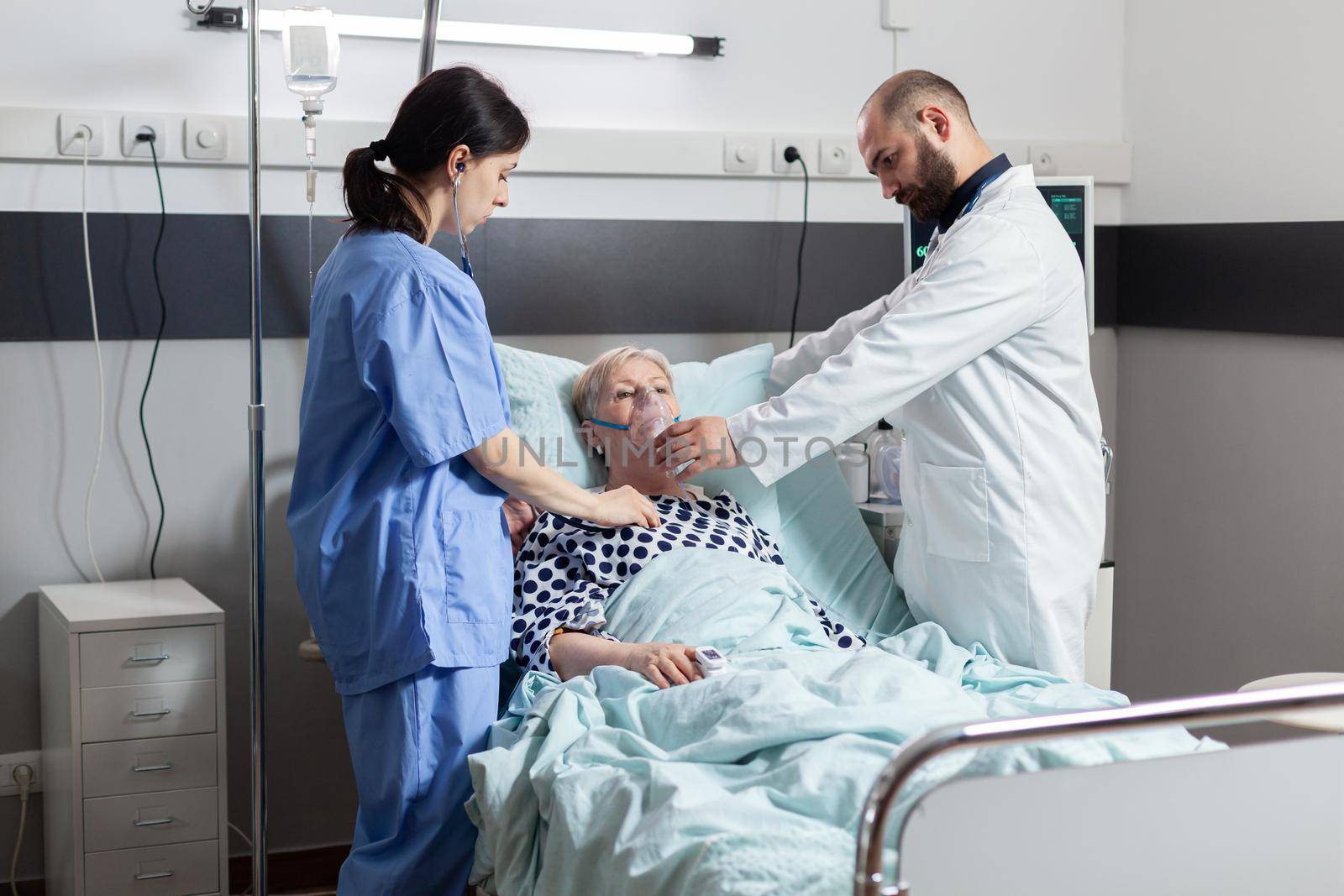 Hospitalized senior woman inhale and exhale through oxygen mask laying in hospital bed. Medical nurse using stethoscope listening patient heart. Eldely woman with oximeter attached on finger.