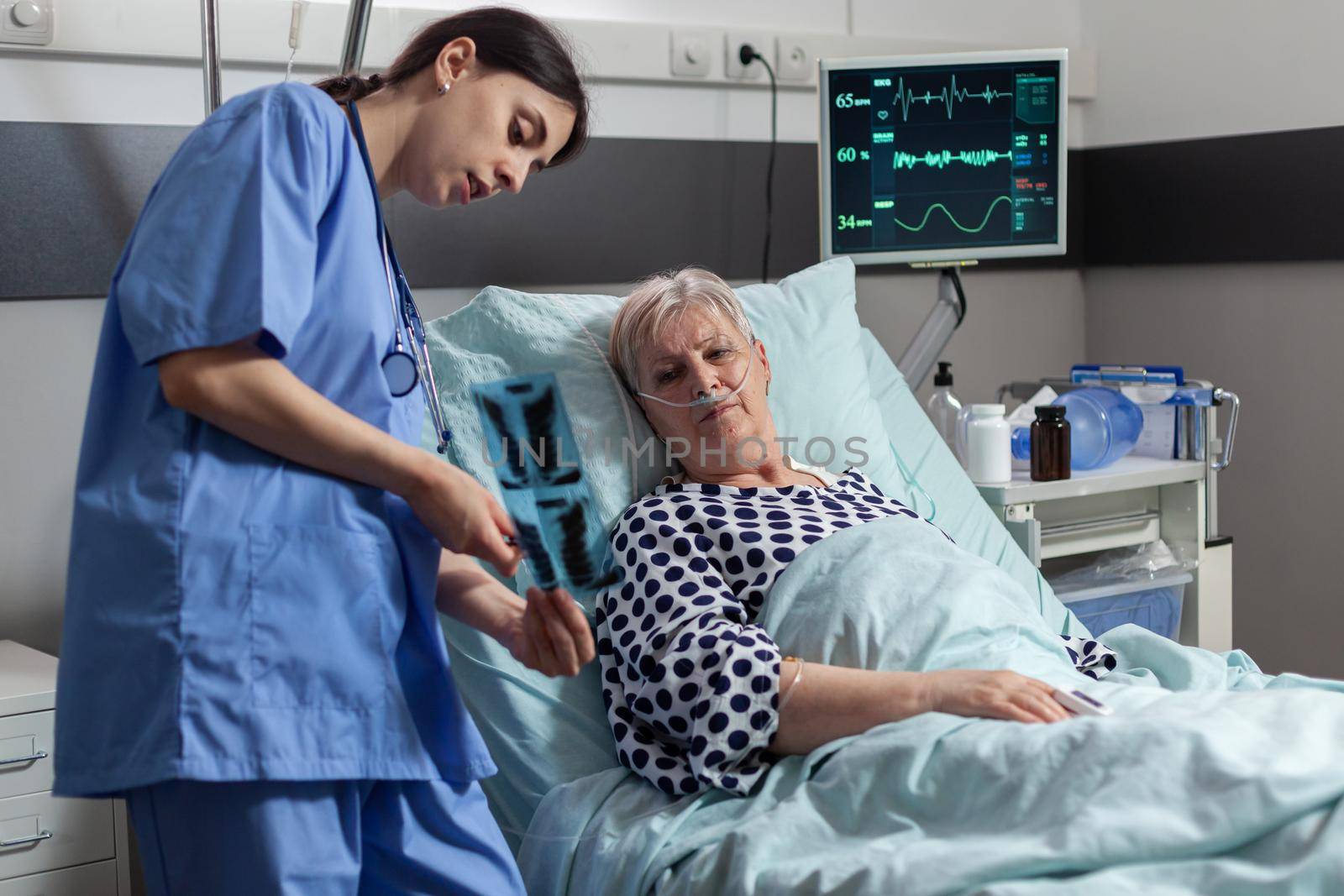 Old patient with lungs illness breathing using oxygen mask laying in hospital bed, listening nurse showing x-ray explayning diagnosis before surgery. Getting treatment through intravenous line.