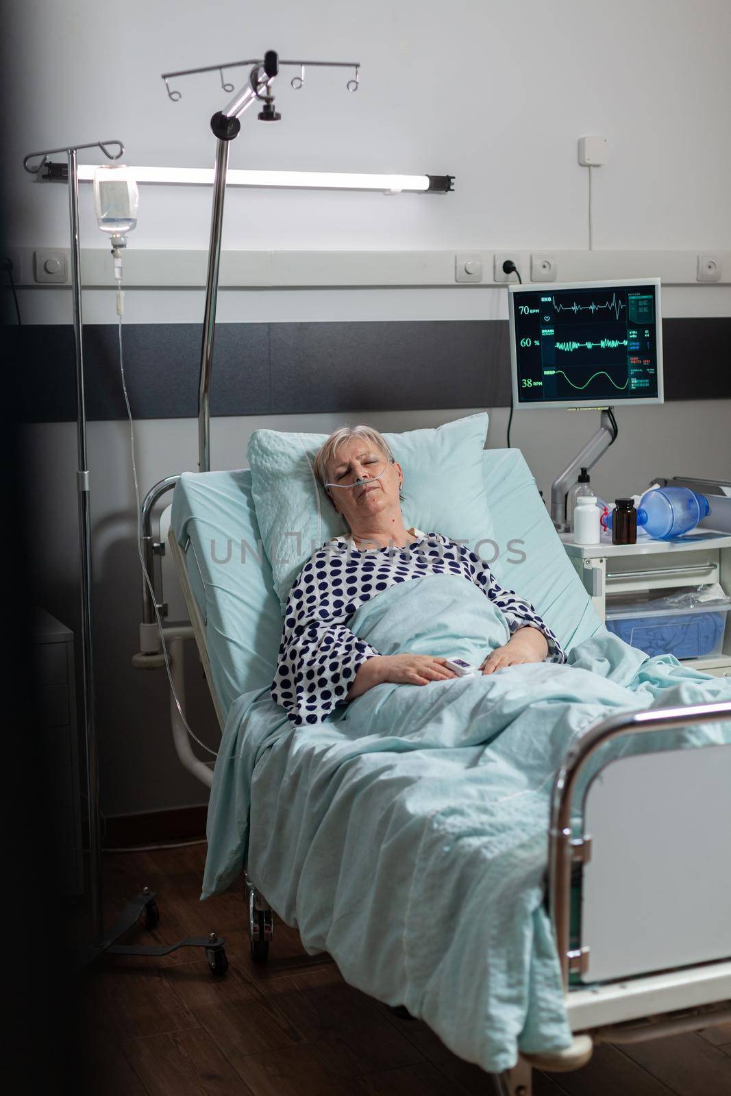 Elderly patient following recovery treatment laying in hospital bed, getting intravenous medicine from iv drip bag. Breathing through oxygen tube because of pulmonary failure.