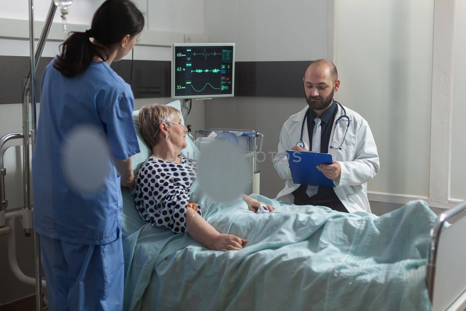 Senior woman patient getting treament through intravenous line and breathing with oxygen tube and oxymeter monitoring level of oxygen saturation. Taking notes on clipboard.