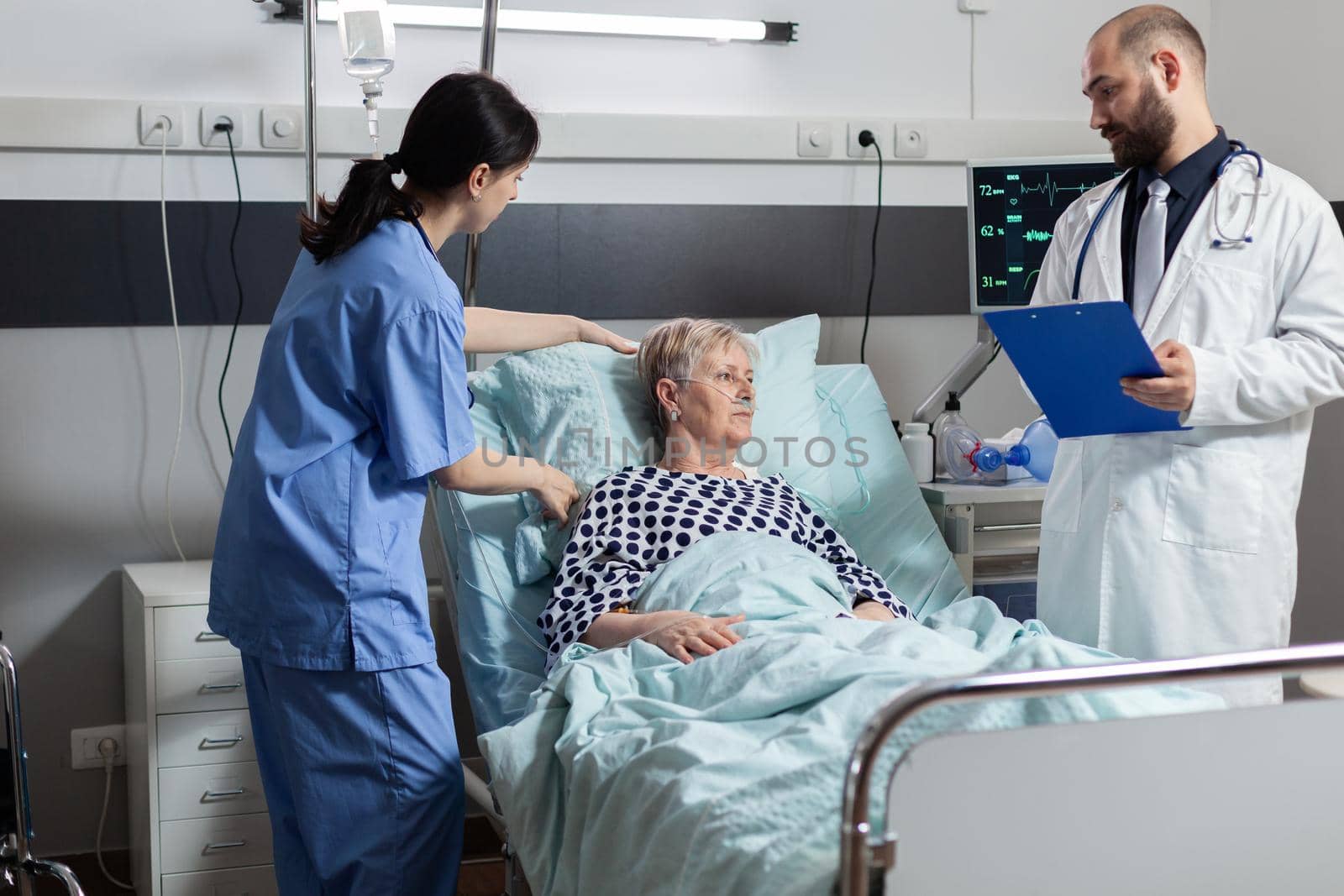 Medical staff checking senior woman health condition in hospital with oxygen tube attached to help her inhale and exhale laying in bed. Nuse making elderly woman comfortable during recovery.