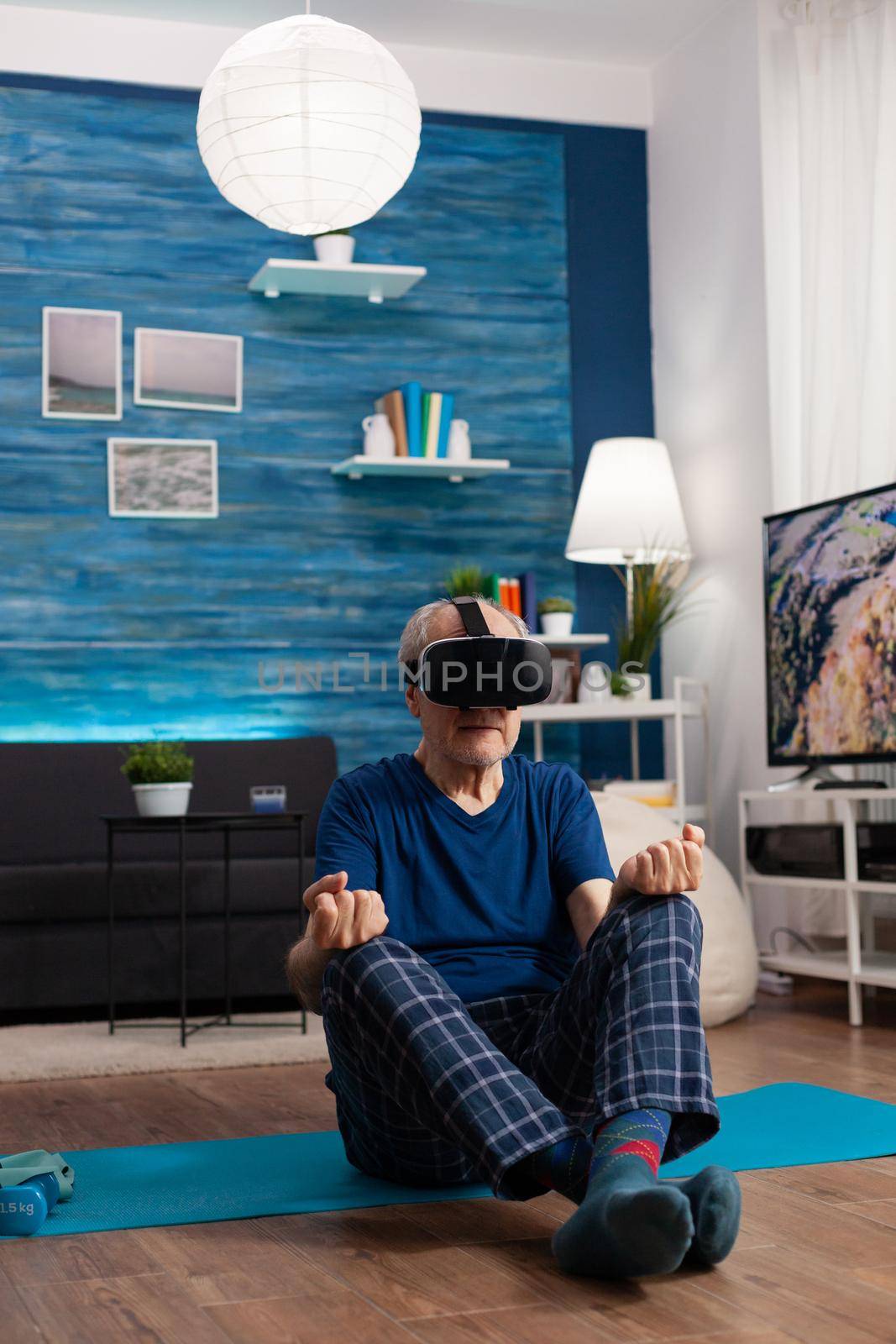 Senior man wearing virtual reality headset meditating while sitting in lotus position on yoga mat in living room during pilates simulation workout. Pensioner training body muscle doing sports exercise