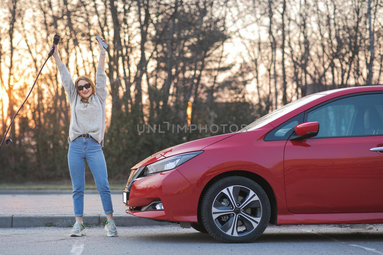 Young adult girl looking at camera having fun and holding in hand power cable supply standing near electric car at charging station