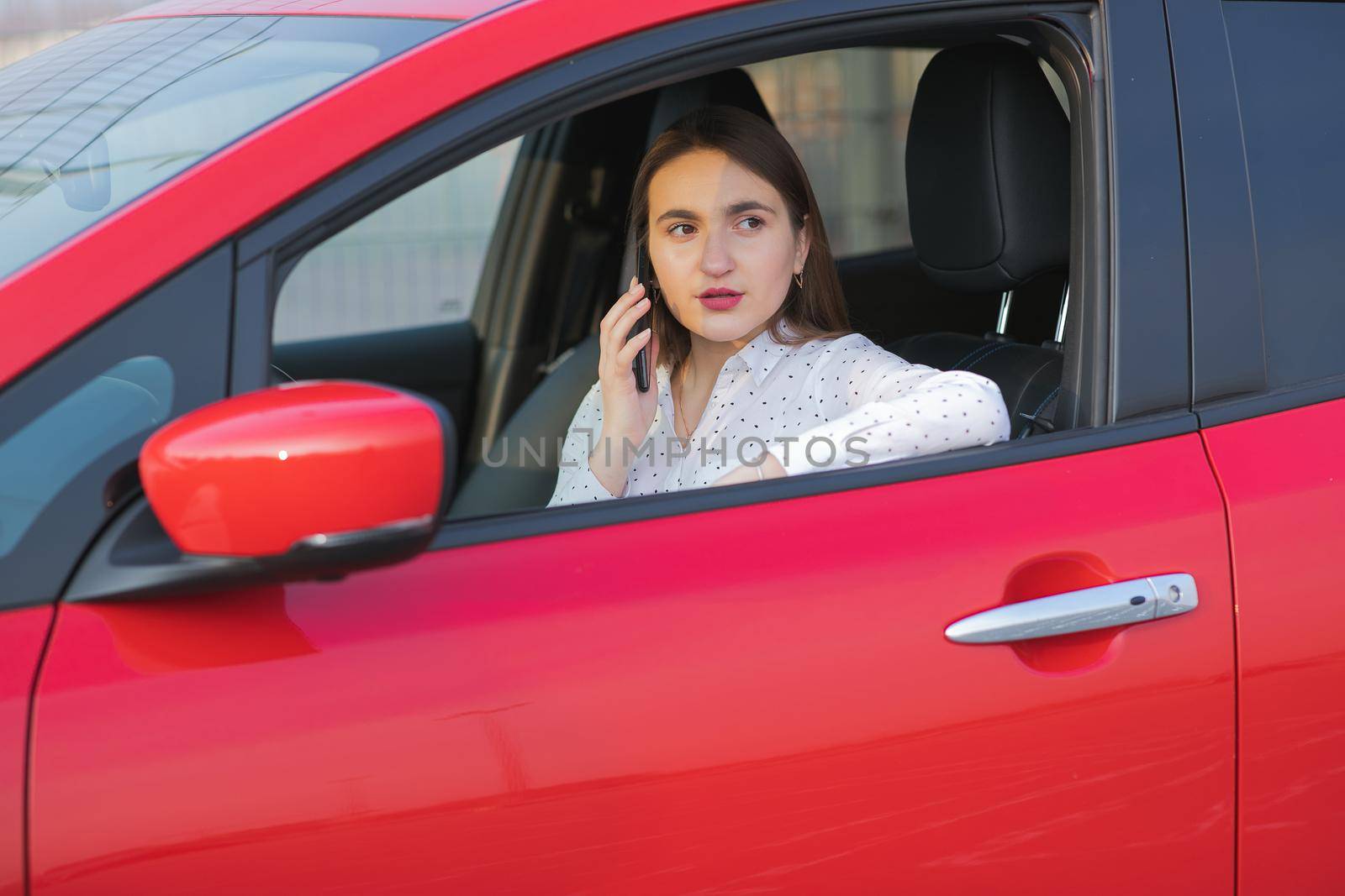 Girl using smart phone and waiting power supply connect to electric vehicles for charging the battery in car. Positive young girl talking on the phone sits in electric car and charging.