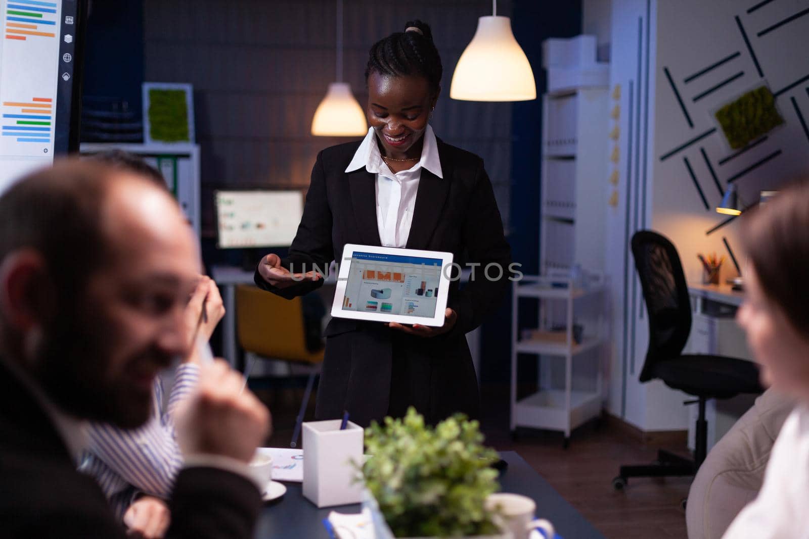 Entrepreneur woman with dark skin overworking in company office meeting room explaining management strategy using tablet. Workaholics multi-ethnic businesspeople brainstorming late at night.