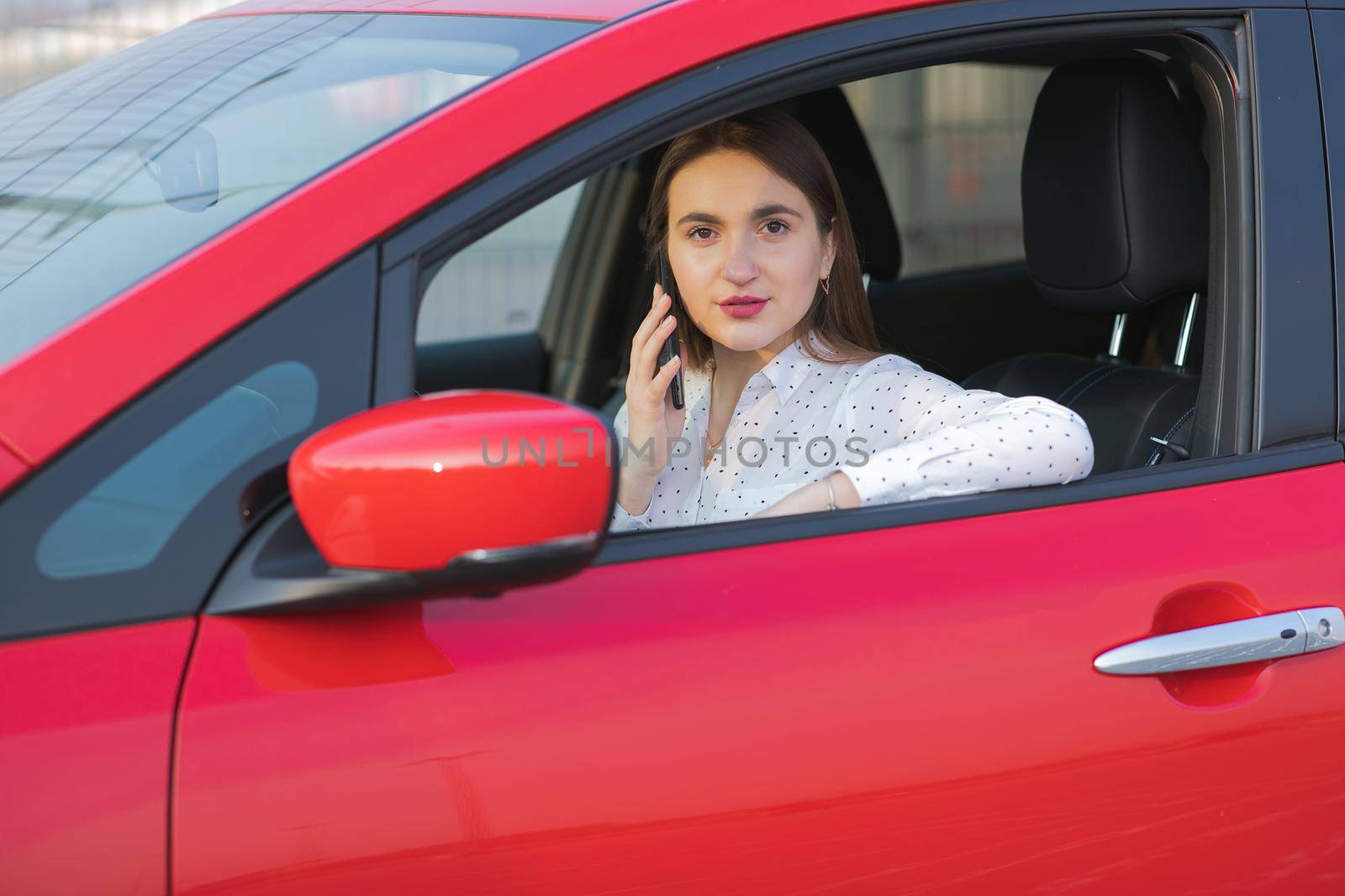 Girl using smart phone and waiting power supply connect to electric vehicles for charging the battery in car. Positive young girl talking on the phone sits in electric car and charging