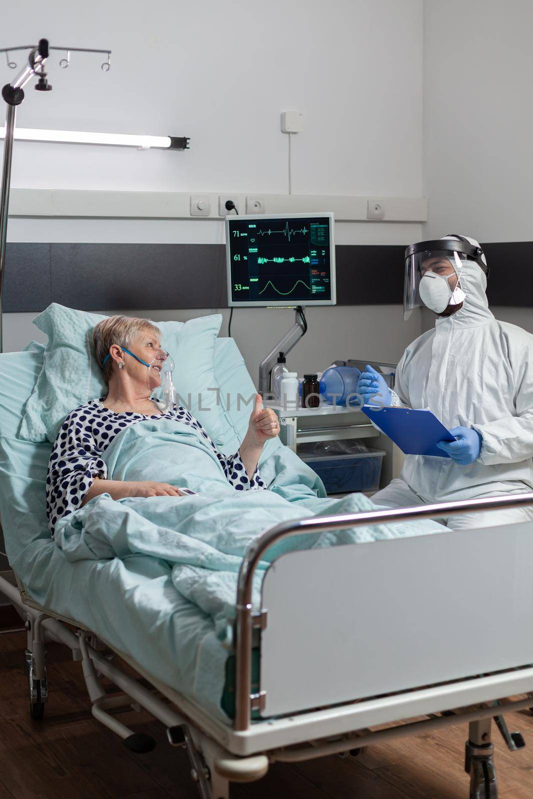 Doctor dressed in ppe suit with face shiled discussing with senior patient, laying in bed with oxygen mask during coronavirus outbreak. Getting intravenous medicine through iv drip bag.