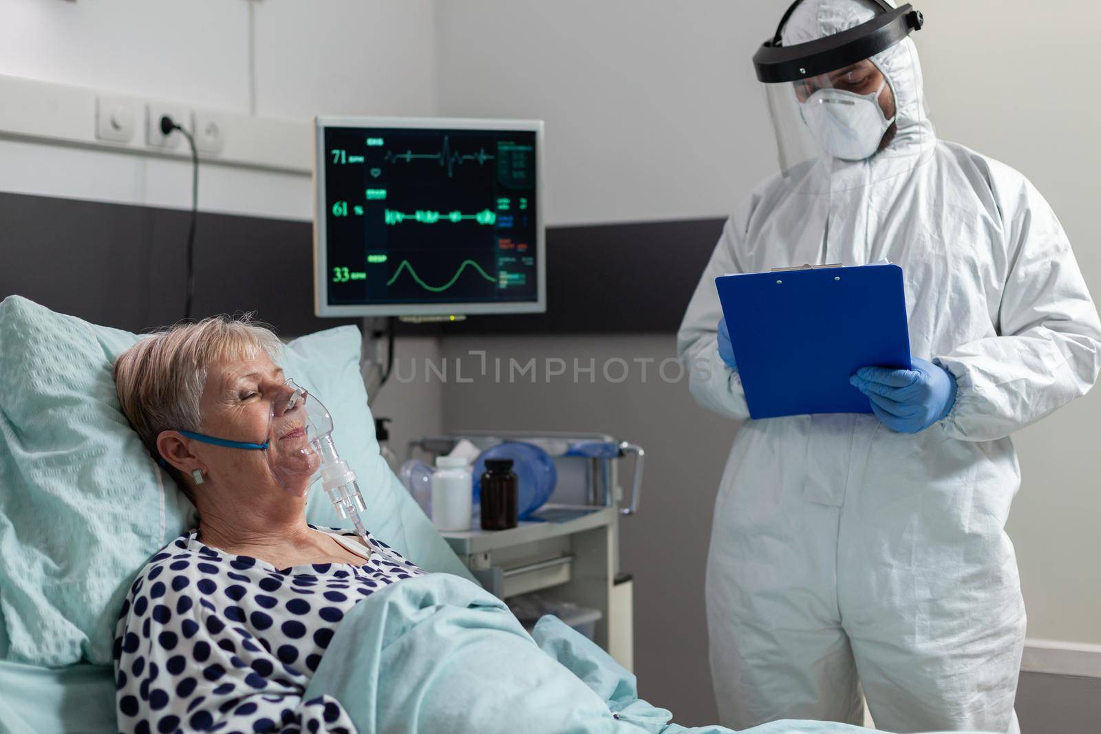 Sick senior patient getting intravenous medicine from iv drip bag laying in bed, inhale and exhale through oxygen mask, during coronavirus pandemic. Doctor wearing ppe suit.