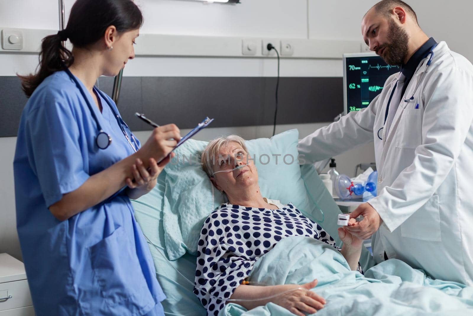 Doctor checking senior patient in hospital intensive care, checking oxygen saturation, blood pressure on oxymeter attached on sick senior patient while nurse making notes on clipboard for diagnosis.