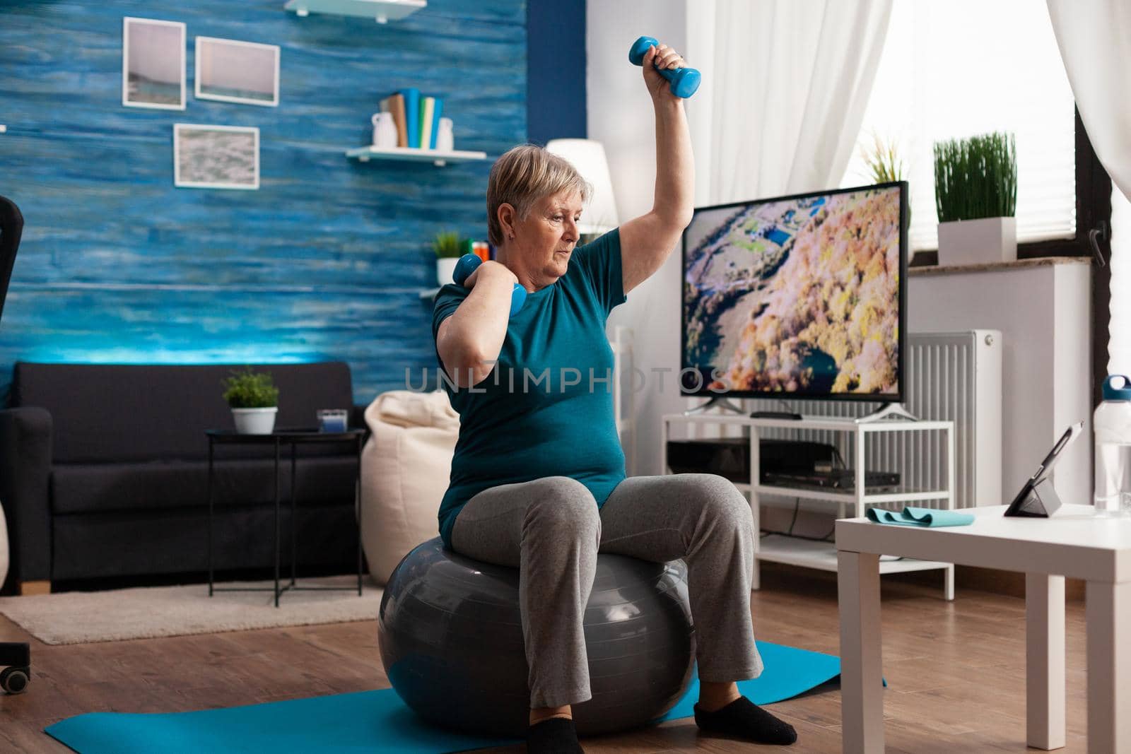 Focused retirement pensioner sitting on fitness swiss ball raising hand streching arm muscle by DCStudio