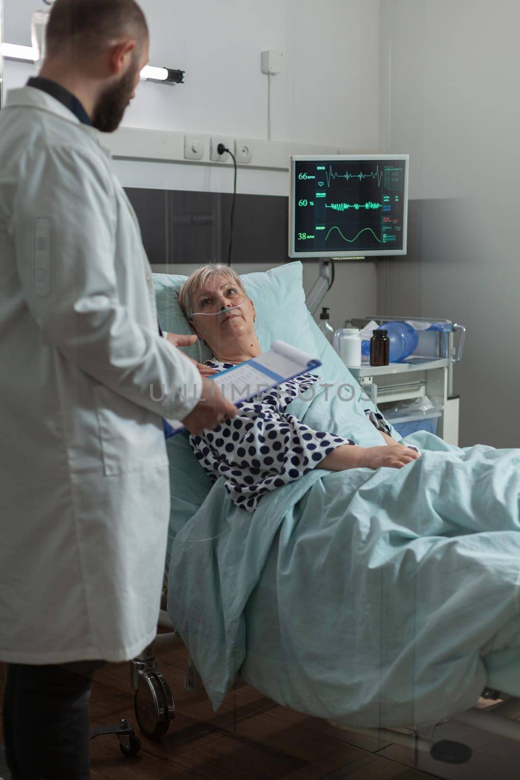 Sick senior woman lying in hospital bed listening doctor talking about treatment and diagnosis. Patient inhale and exhale with help from oxygen tube. Iv drip bag attached on arm.